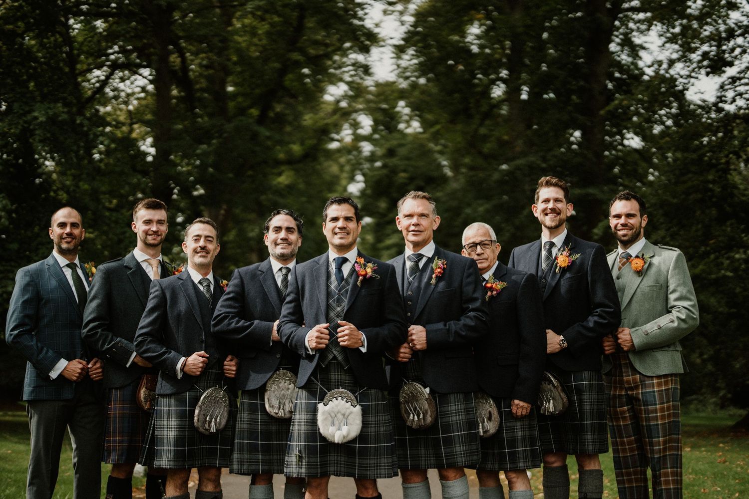 Wedding party in kilts and formal attire pose together on a green lawn.