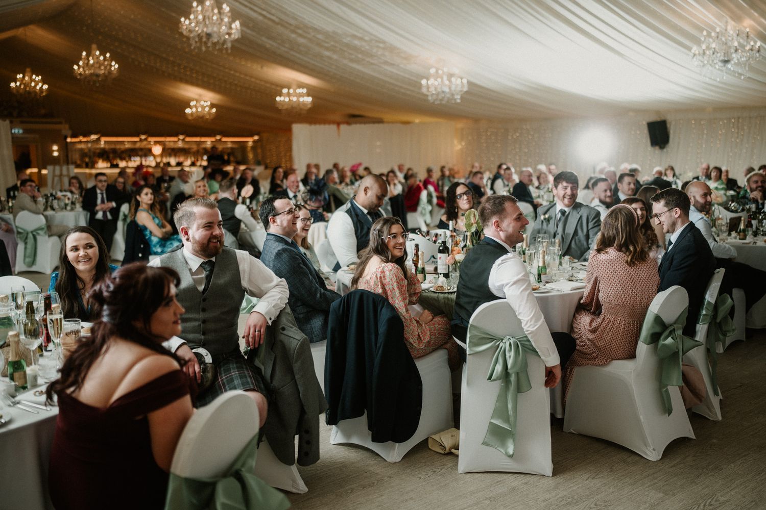 Large wedding reception hall filled with guests at round tables under crystal chandeliers.