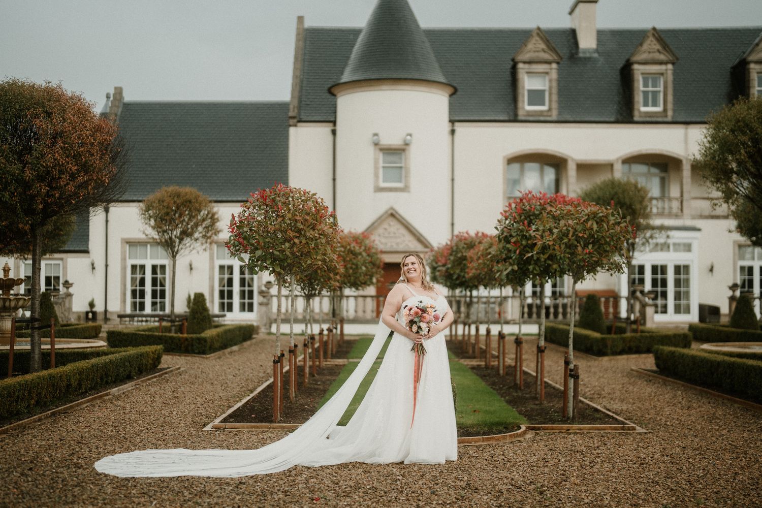 A wedding photo in front of a grand white mansion with manicured gardens and gravel paths.