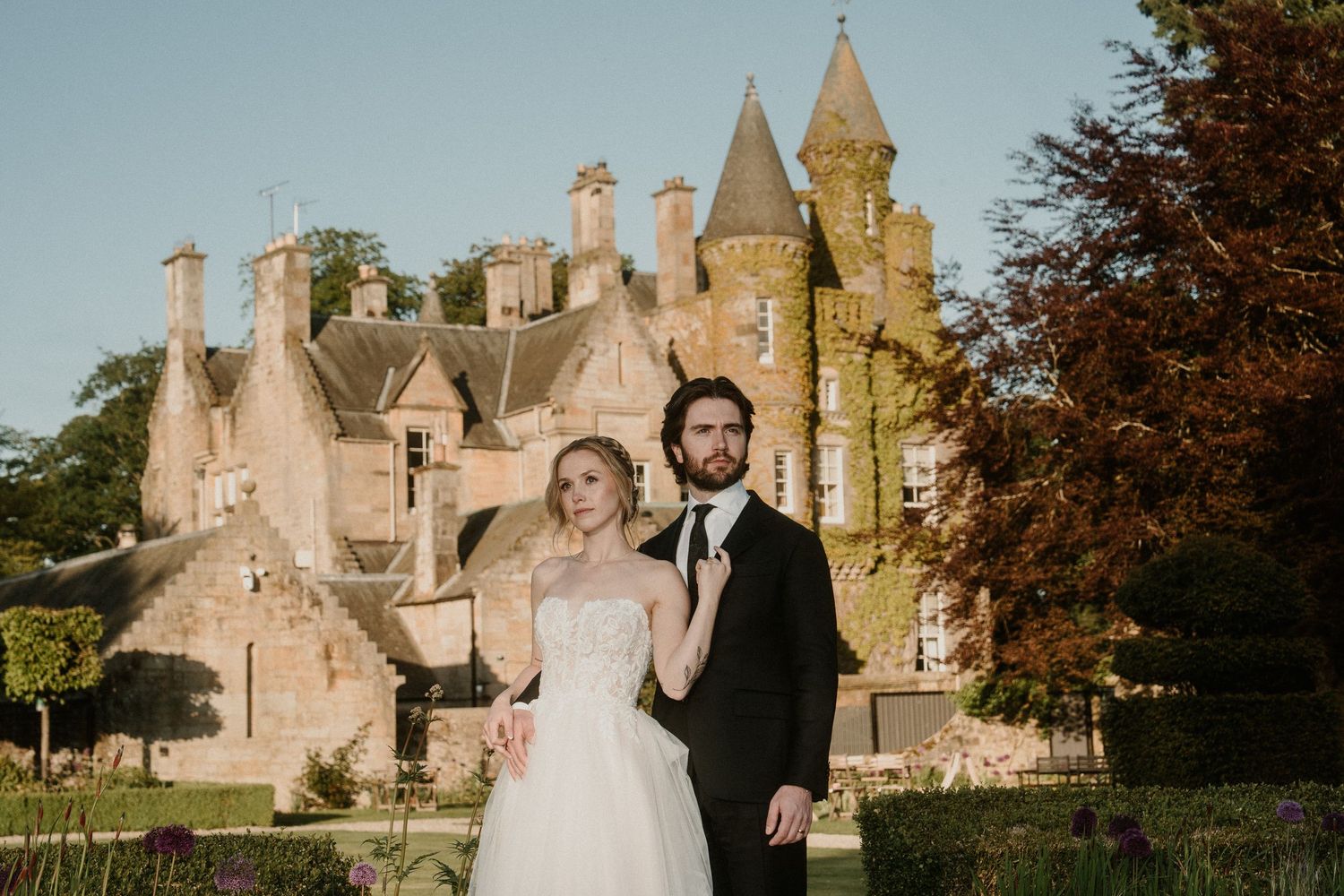 Wedding couple stands in front of a majestic stone castle with Gothic architecture and spires.