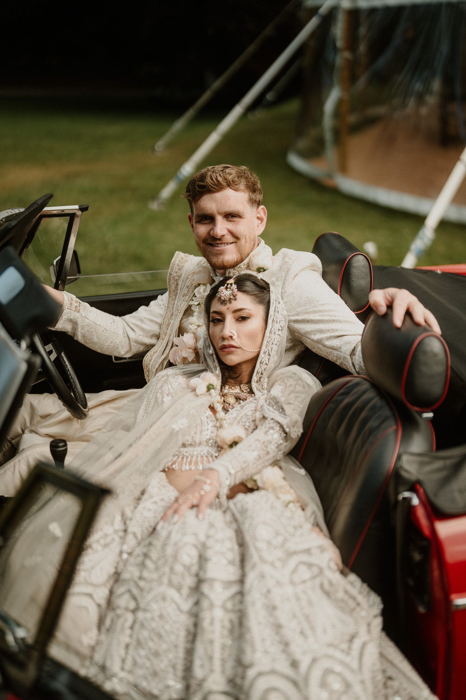 Romantic couple poses in vintage red convertible car during wedding photoshoot.