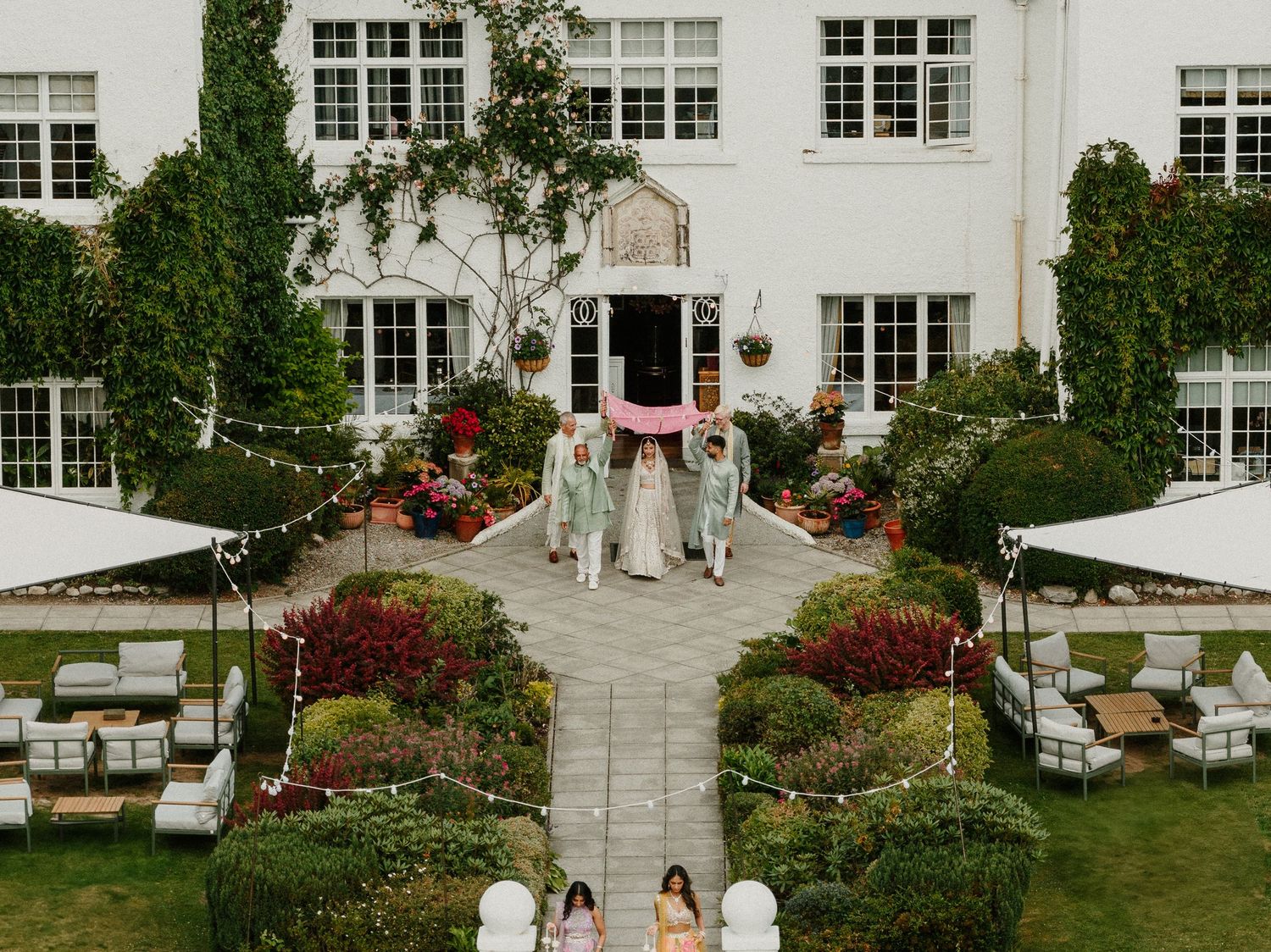 Elegant white mansion with manicured gardens serves as backdrop for outdoor wedding ceremony.