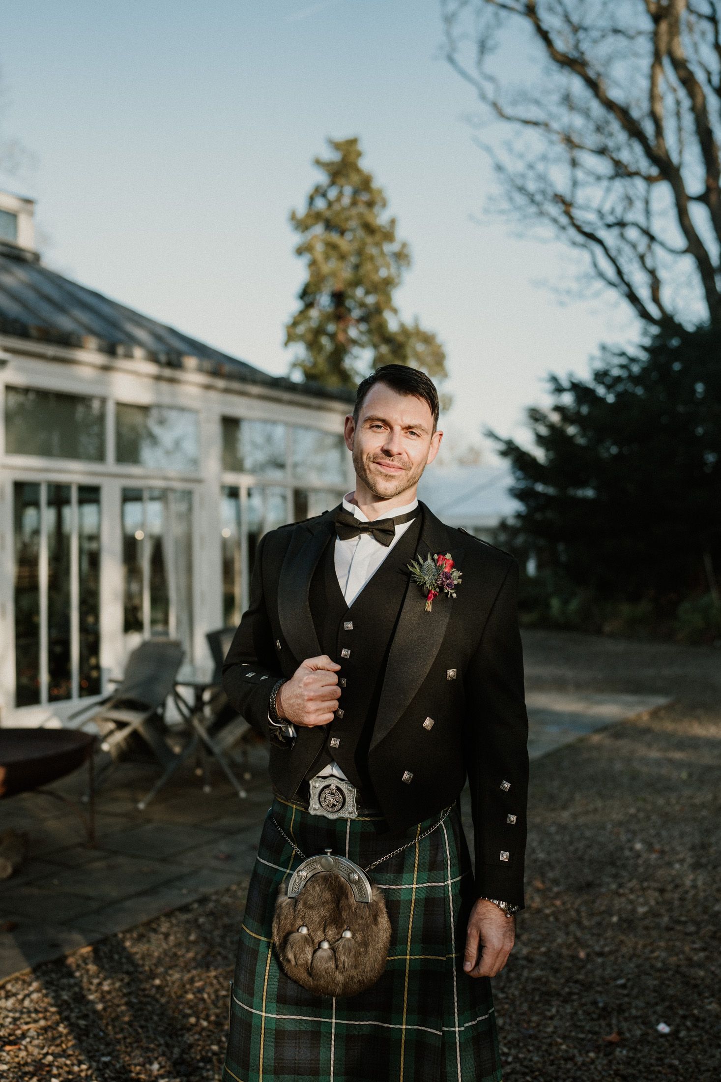 A groom in traditional Scottish formal wear with green tartan kilt poses outside a conservatory.