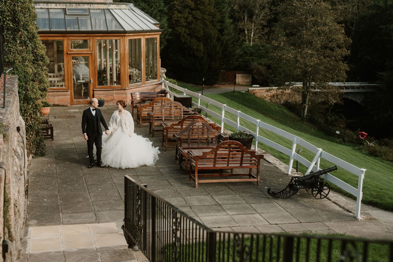 Wedding couple walking along stone building exterior with wooden benches.