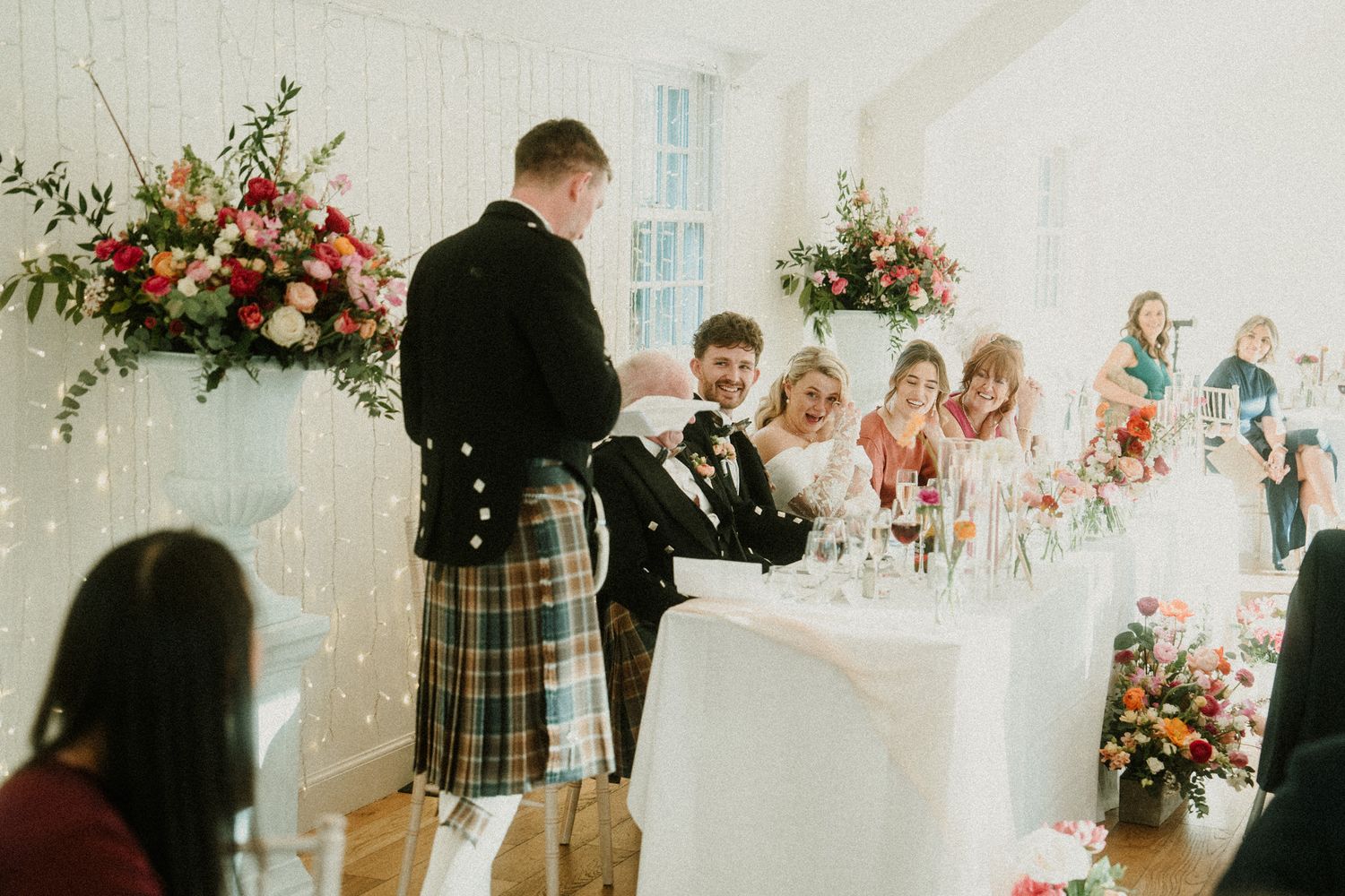 Wedding reception venue with floral centerpieces and white table settings in a bright room.