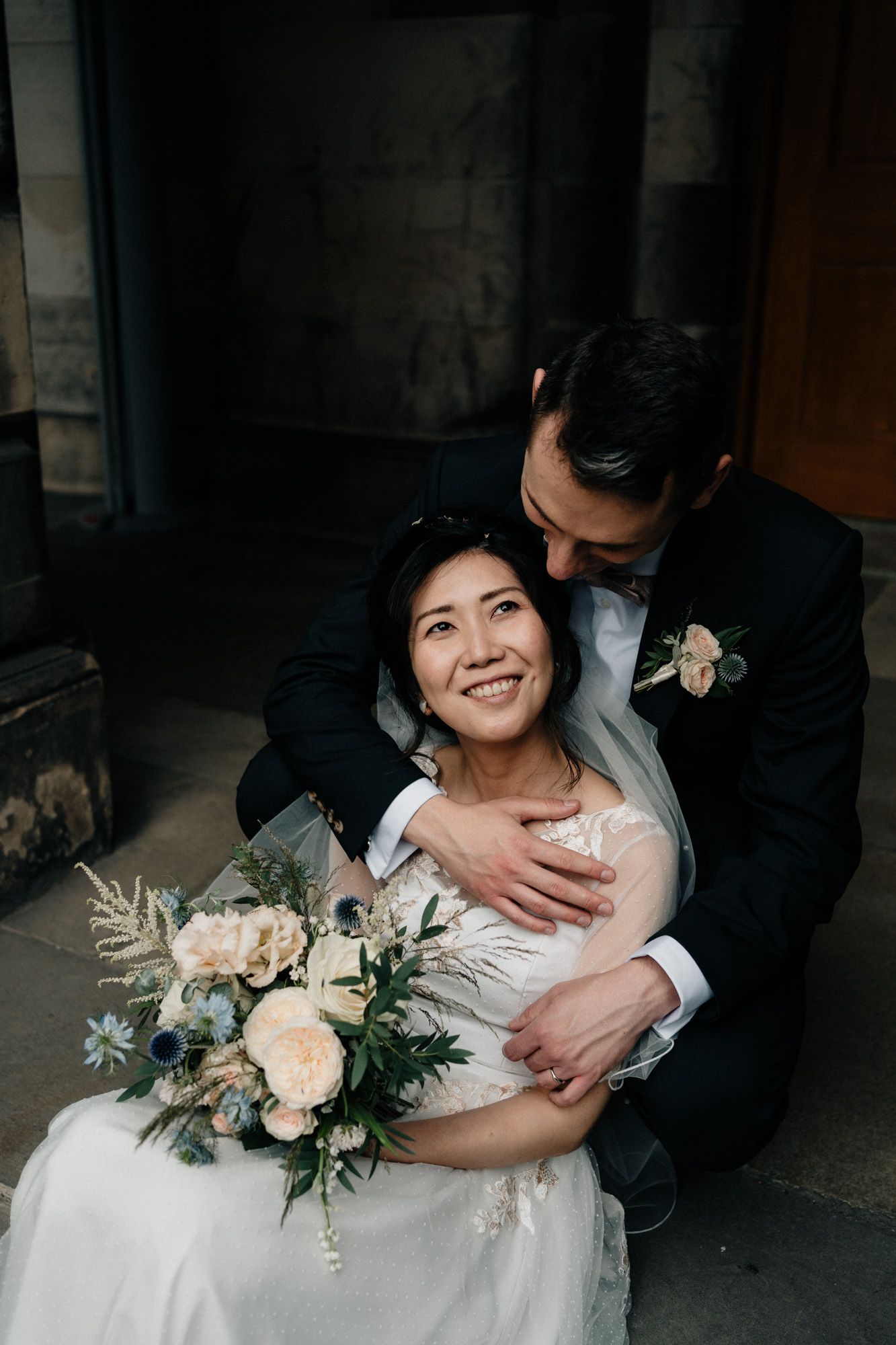 Wedding couple embracing with elegant floral bouquet featuring peach roses and greenery.