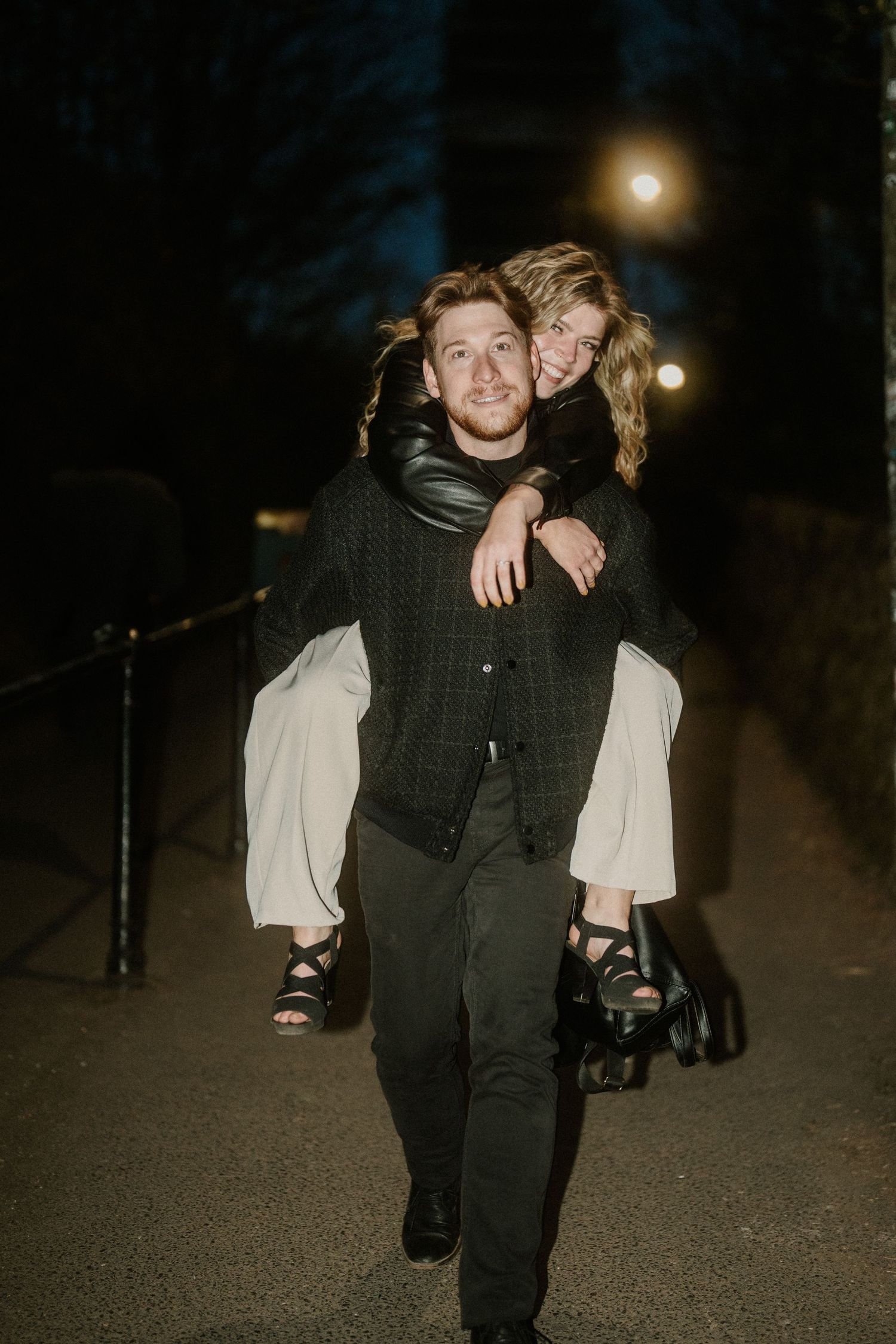 A man gives a smiling woman a piggyback ride at night on a lit pathway, both dressed in dark elegant attire.