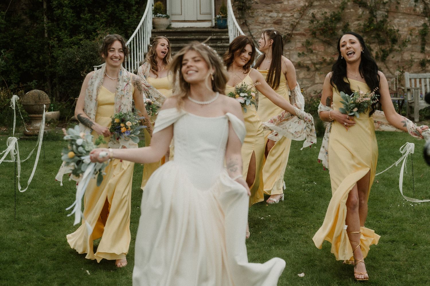 Bride in white gown laughs joyfully with bridesmaids in yellow dresses running across green garden lawn.