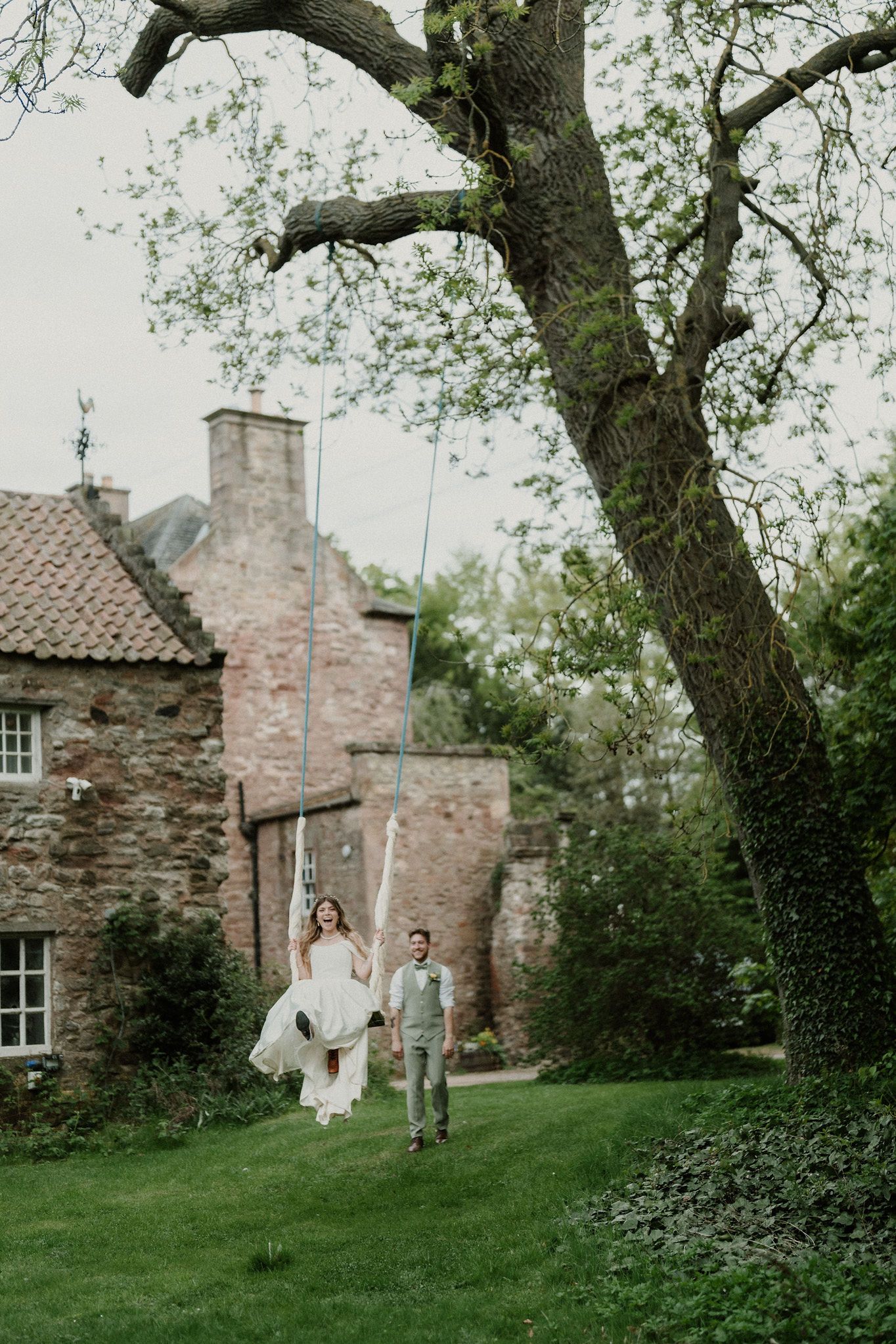 Bride on tree swing held by groom in lush garden of rustic stone wedding venue.