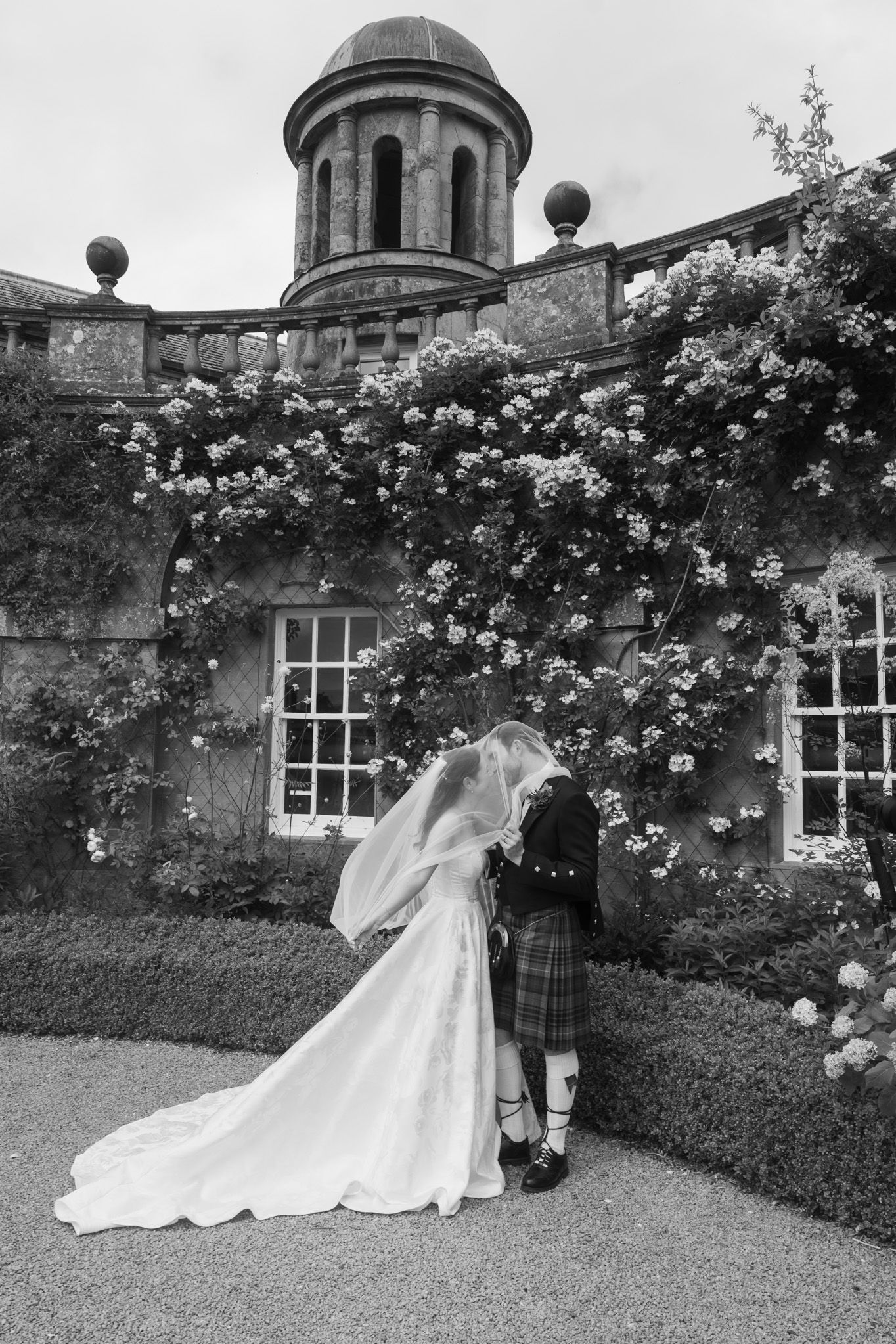 Black and white photo of bride and groom kissing in a Scottish garden, groom in kilt, bride in flowing gown with veil.