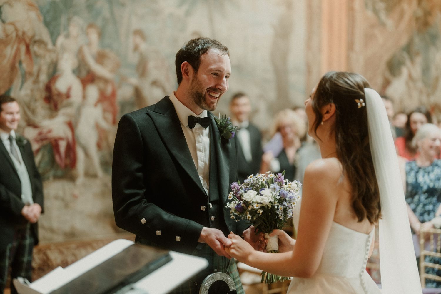 Groom in Scottish kilt laughing joyfully while exchanging vows with bride holding a purple bouquet at altar.