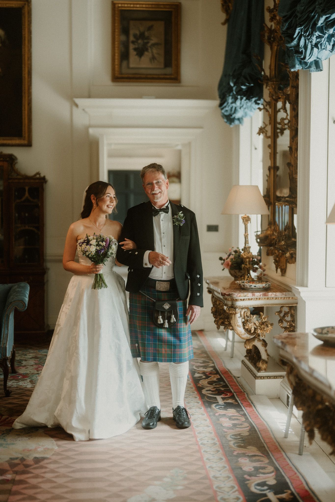Bride in white gown holding bouquet walks with groom in traditional Scottish kilt inside elegant wedding venue.