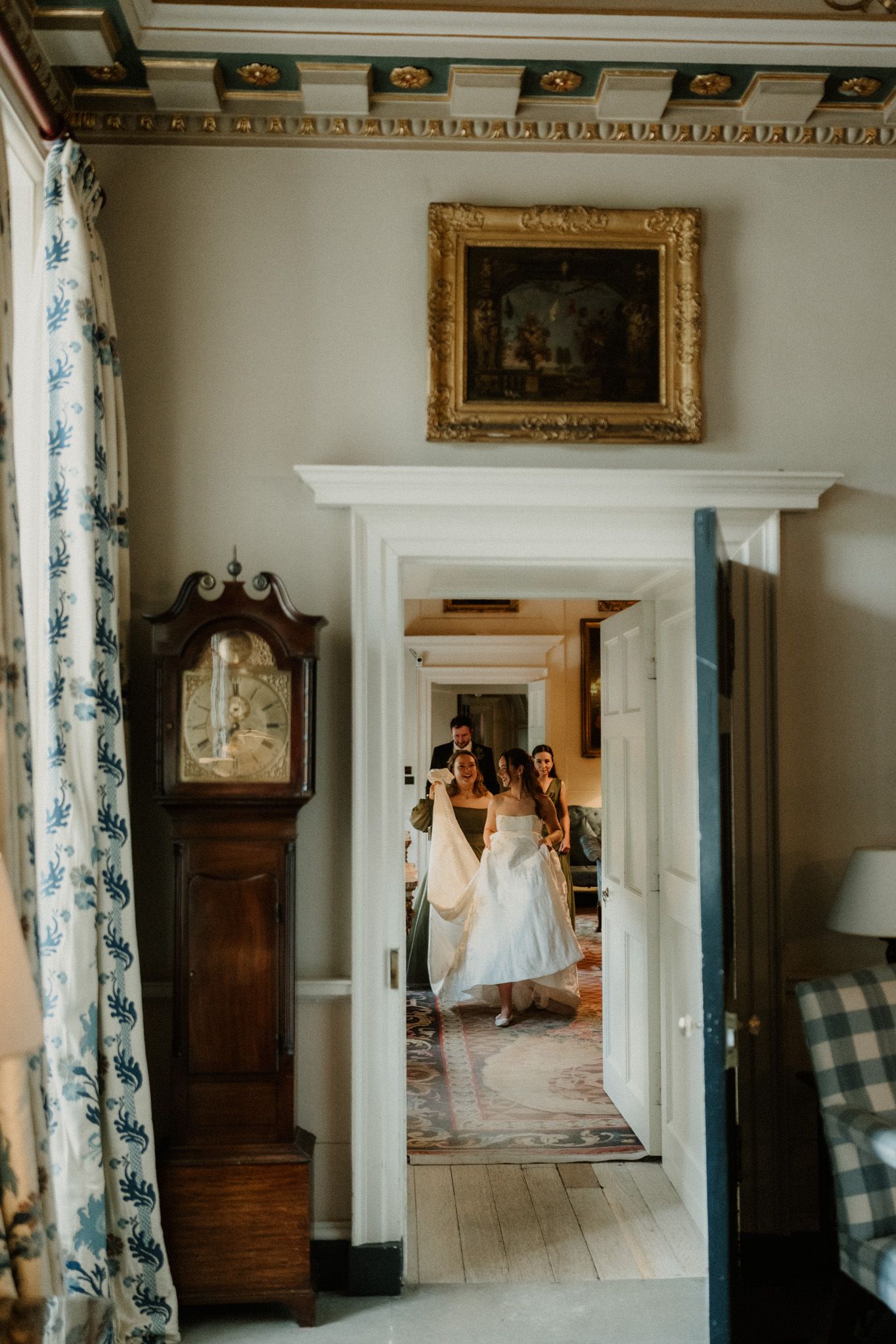 Bride in white dress walks through elegant hallway with grandfather clock and ornate gold framed painting.