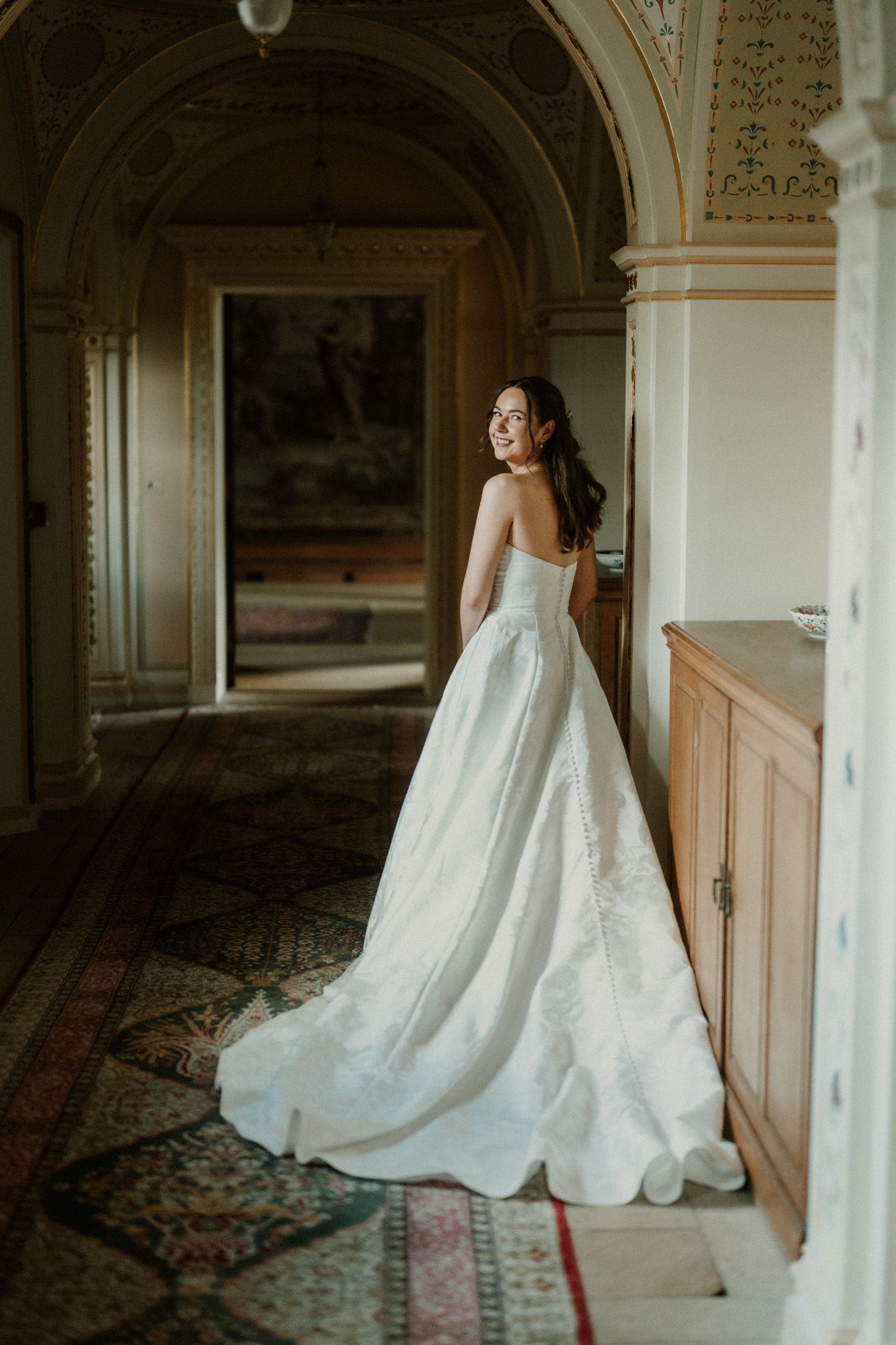 Bride in a strapless white ball gown with a long train stands in an elegant hallway, smiling over her shoulder.