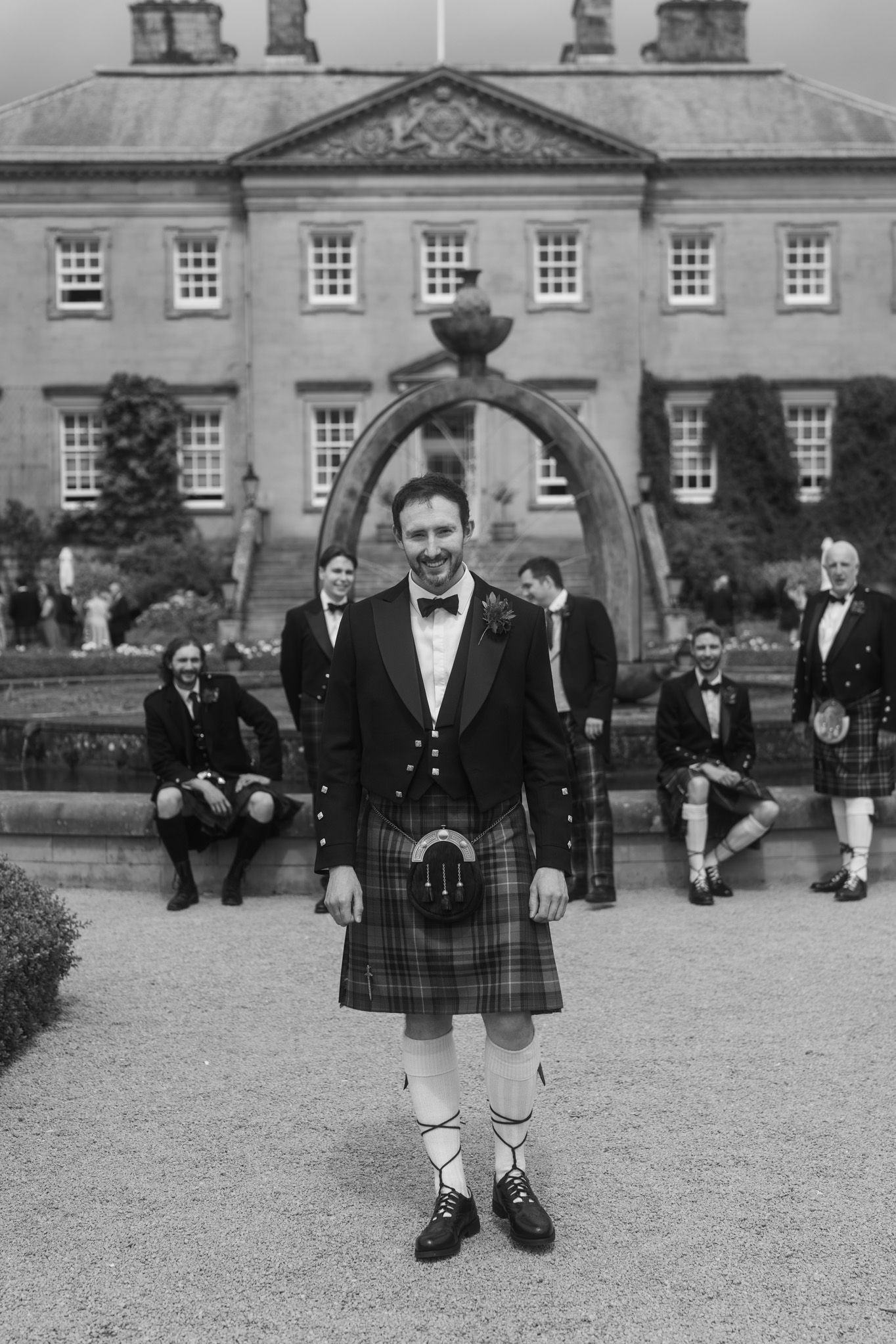 Groom in traditional Scottish kilt stands before a grand estate with groomsmen relaxing behind him.