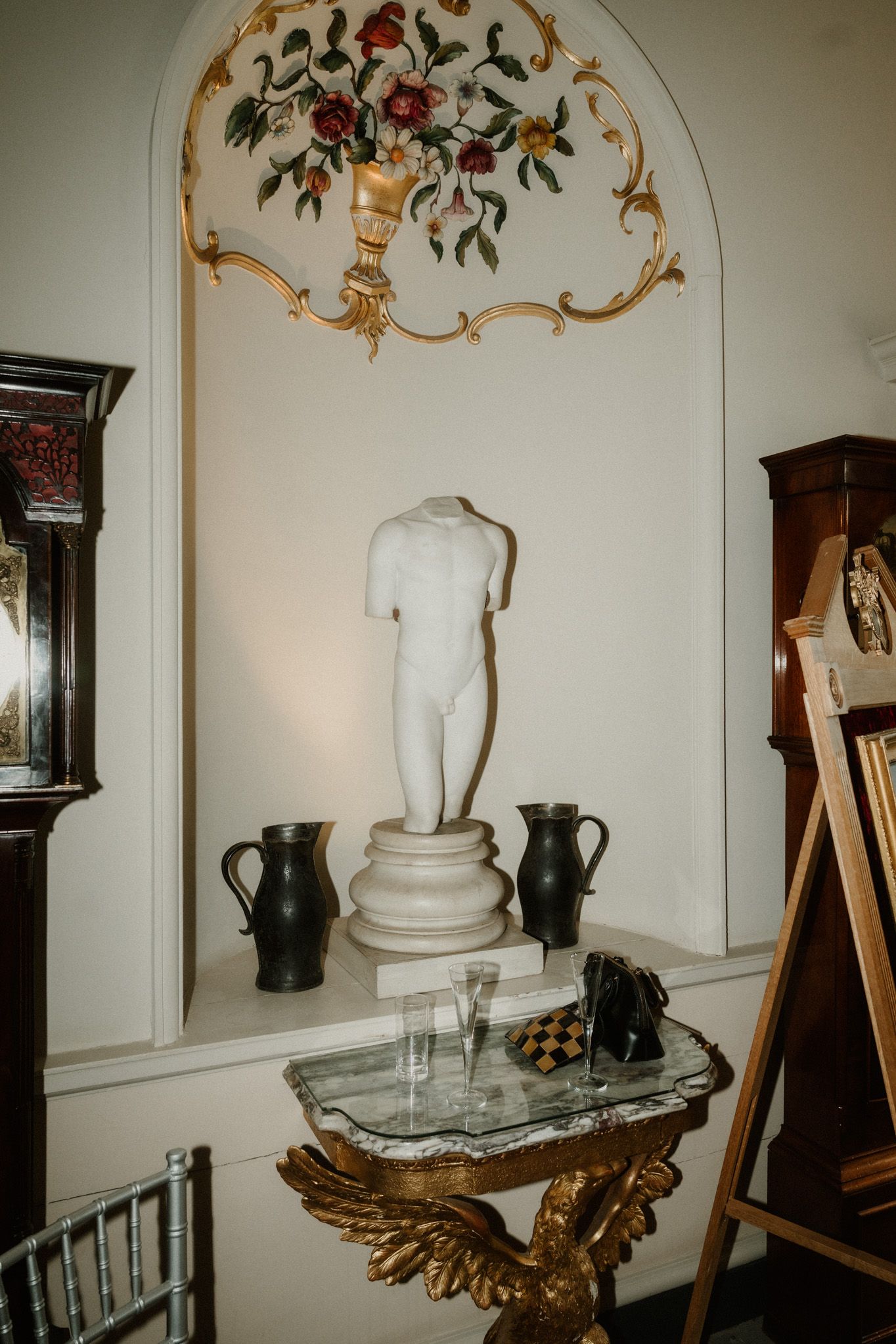 Classical marble torso sculpture displayed on pedestal with decorative floral gilt mirror and antique furnishings in elegant interior.