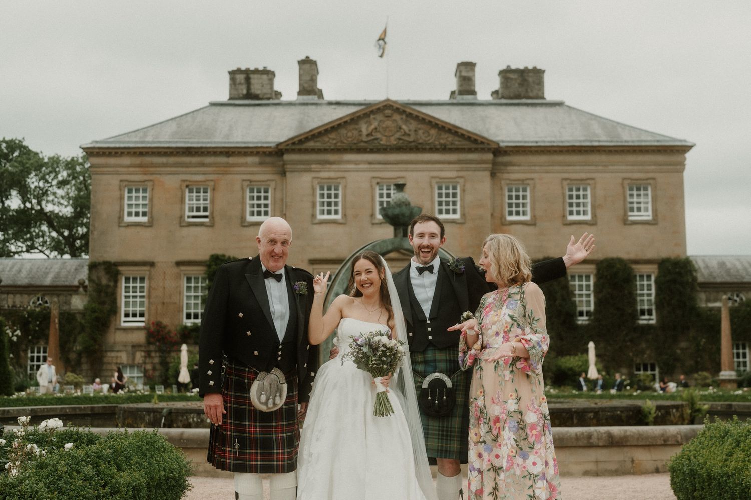 Wedding party posing in front of grand Scottish estate, bride in white gown, groom and man in traditional kilt.
