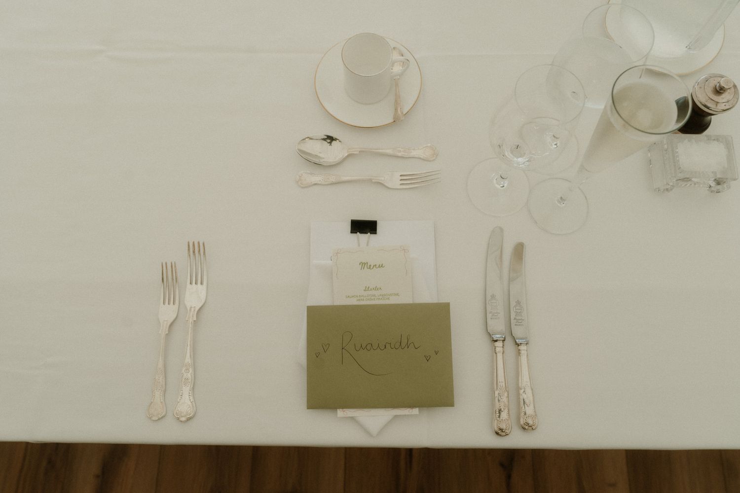 Elegant wedding place setting with gold menu card, silverware, white espresso cup, and crystal glasses on white tablecloth.