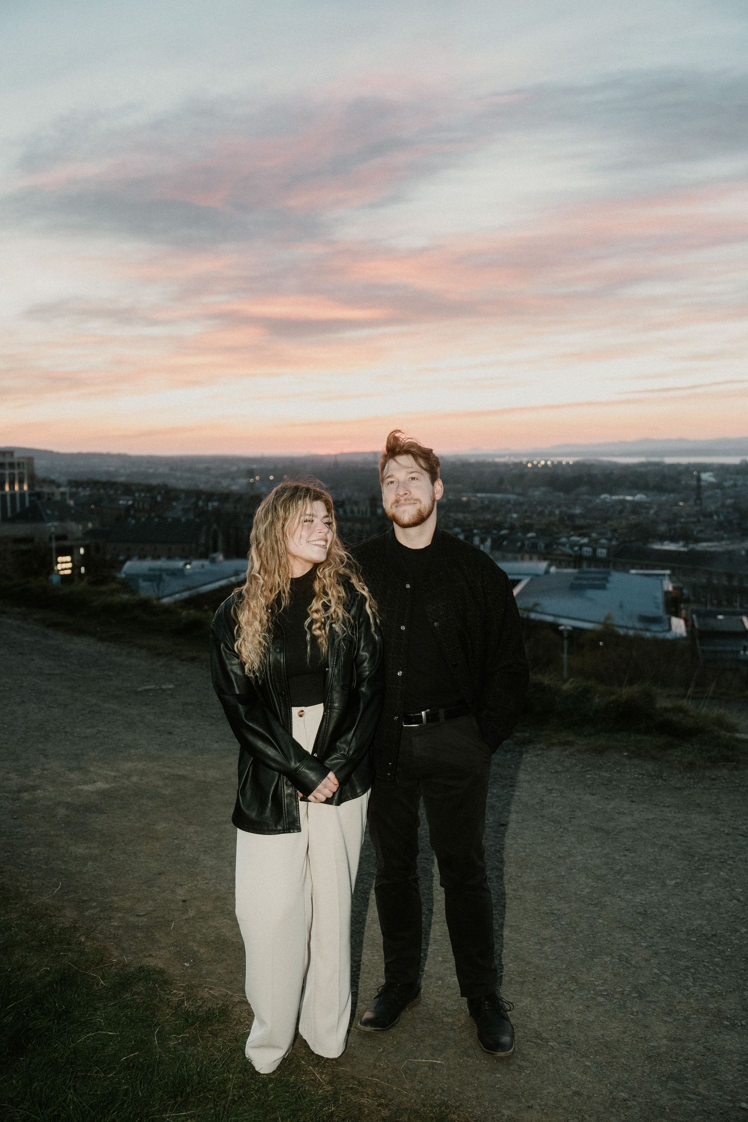 Couple standing together on a hilltop at sunset with a city skyline and colorful pink sky in the background.