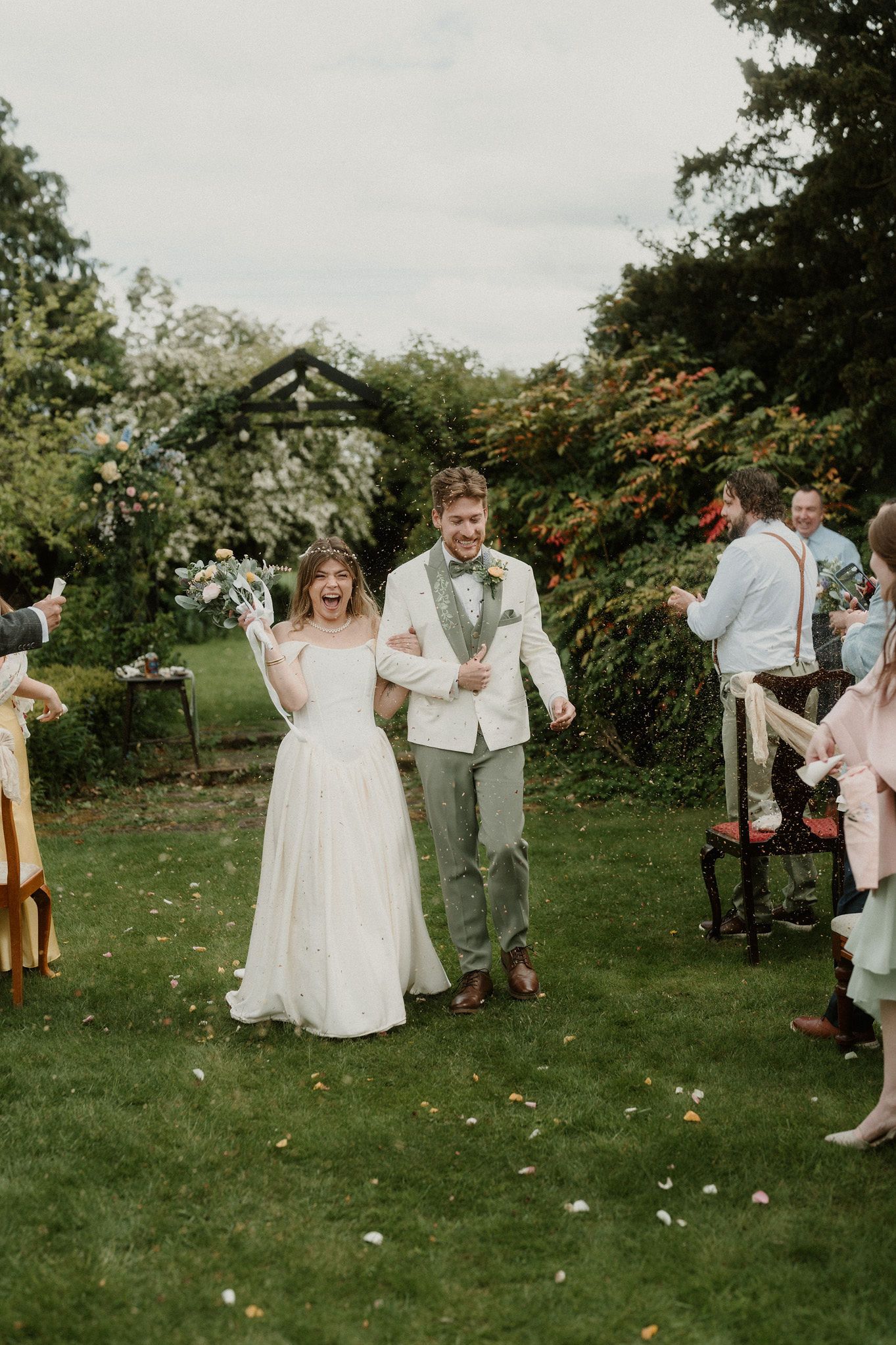 Joyful bride and groom walk through confetti shower at outdoor garden wedding ceremony surrounded by cheering guests.