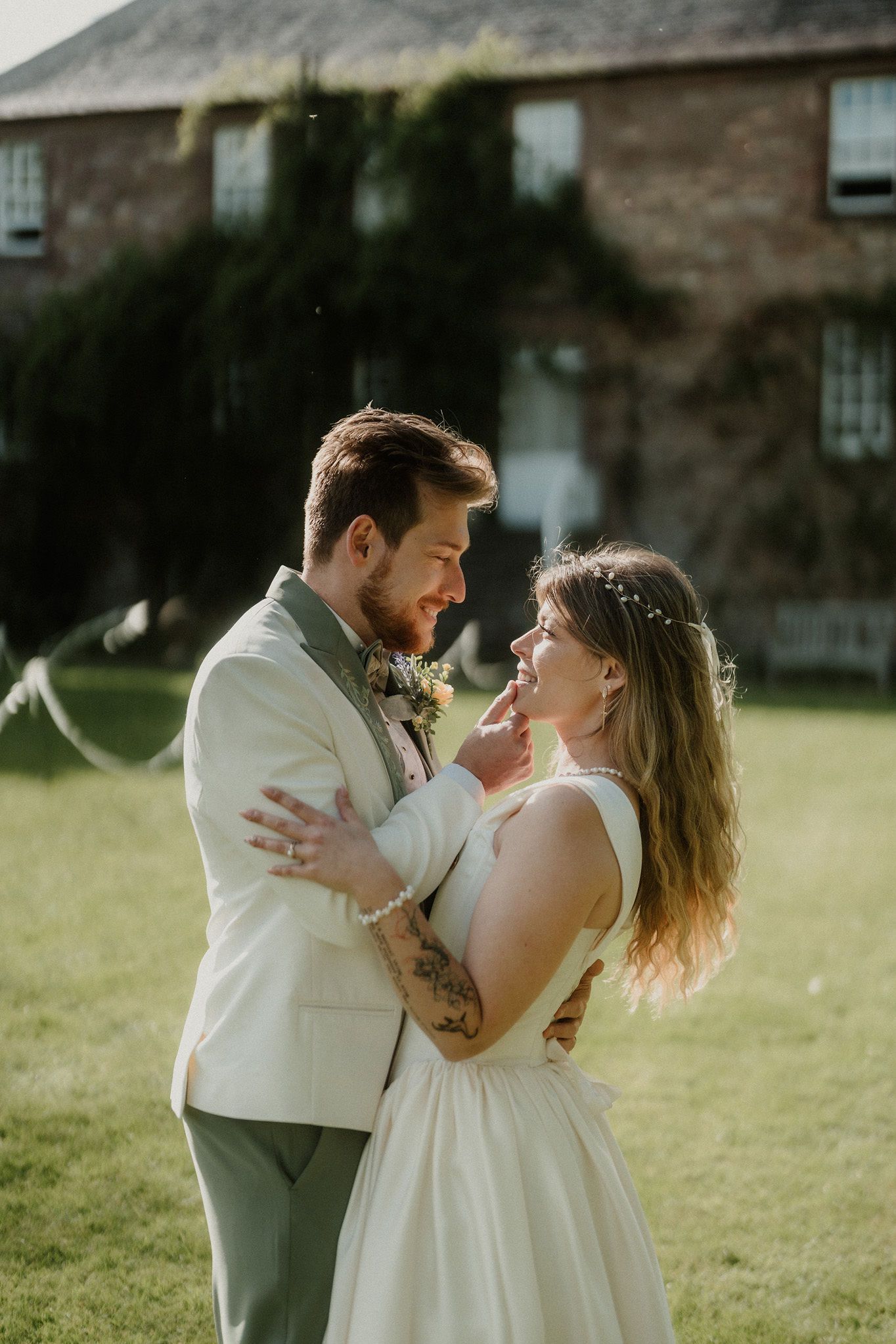 Newlyweds share romantic embrace on sunlit lawn with tattooed groom in sage suit and bride.
