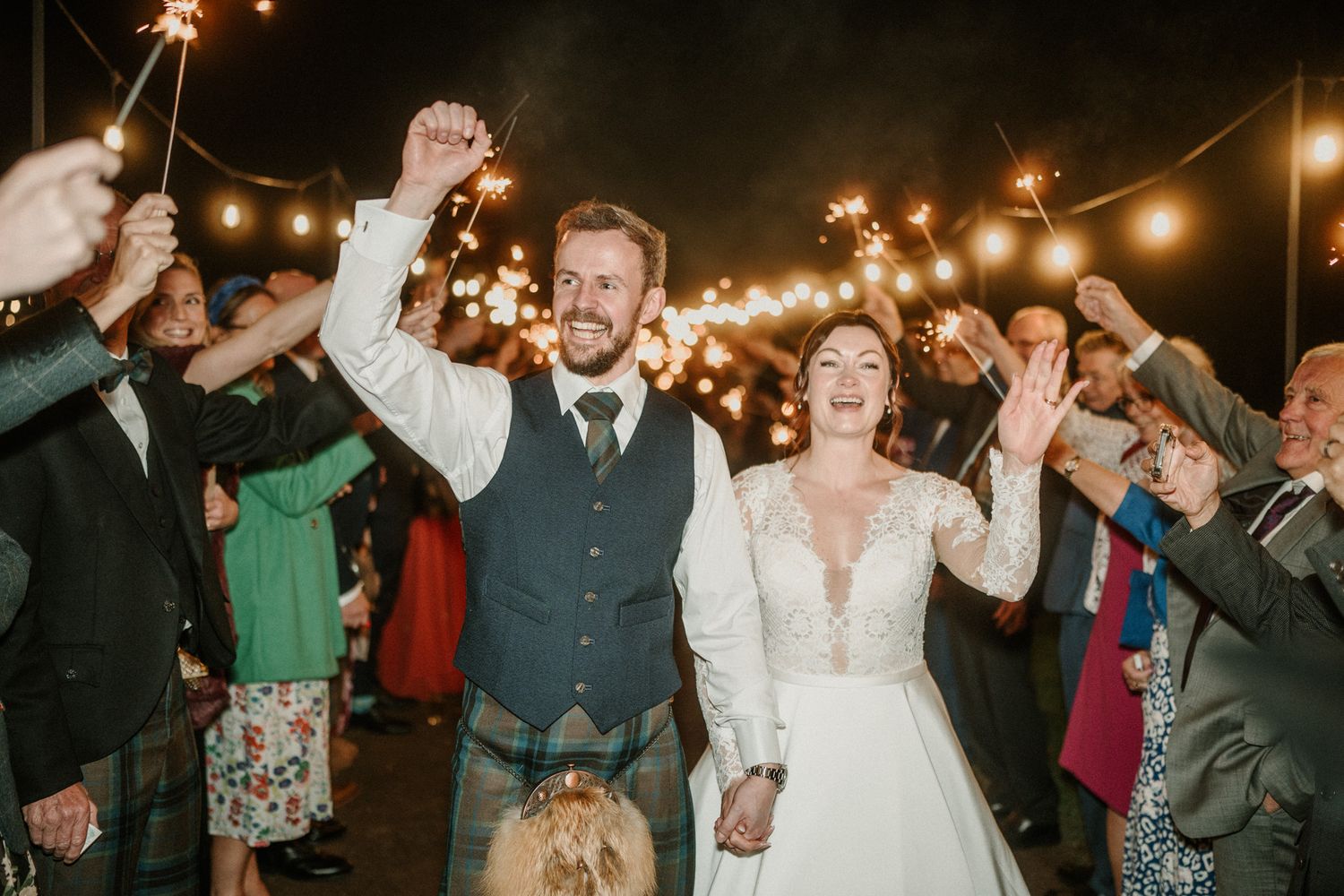 Sparklers illuminate a joyous wedding celebration as guests create a glowing tunnel outdoors.