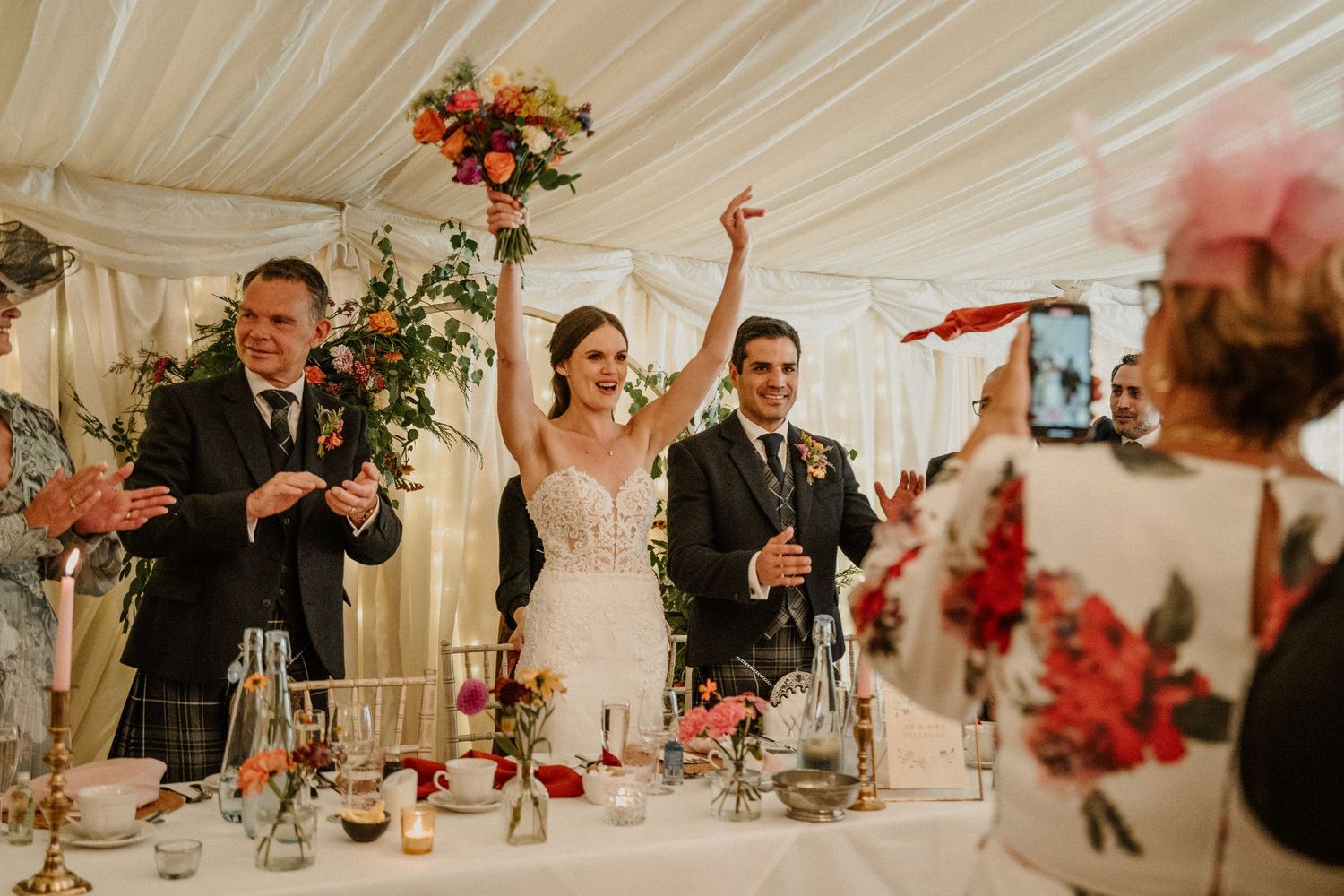 Elegant wedding reception in a white marquee tent with red floral centerpieces and draped fabric ceiling.