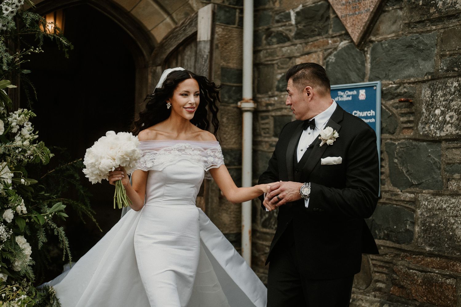 Elegant wedding couple poses against stone wall with romantic lighting at dusk.