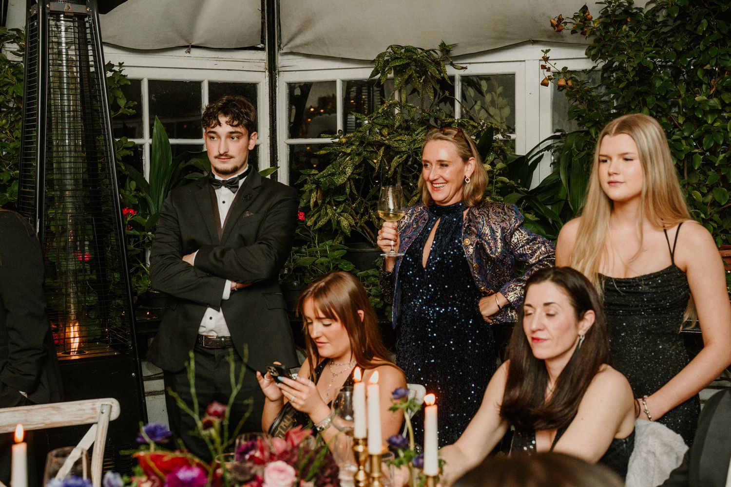 Wedding guests seated at long tables in a glass-enclosed venue with lush greenery.