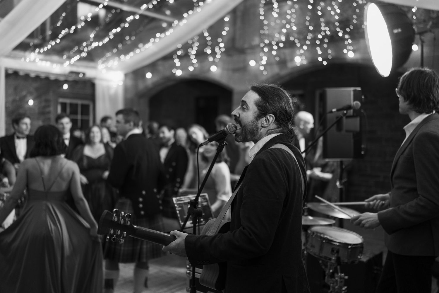 Live band performs at an evening wedding reception as a guitarist sings into a microphone while guests mingle under string lights.