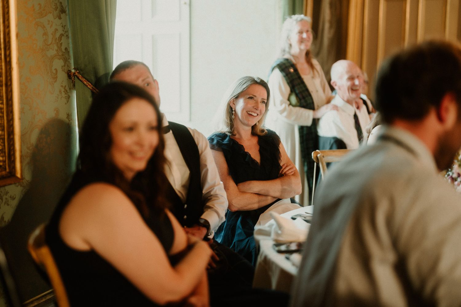 Smiling wedding guests seated at a table laugh and enjoy a speech in an elegant room with green drapes and warm natural light.