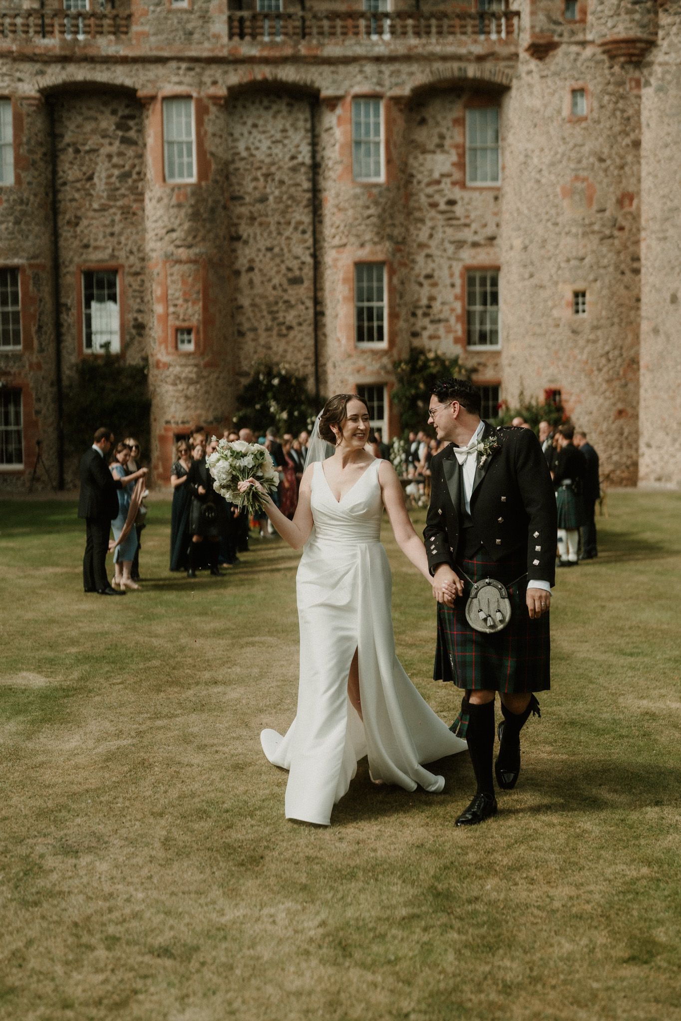 Bride in a white gown and groom in a kilt hold hands and walk across a lawn before wedding guests and a historic stone castle.