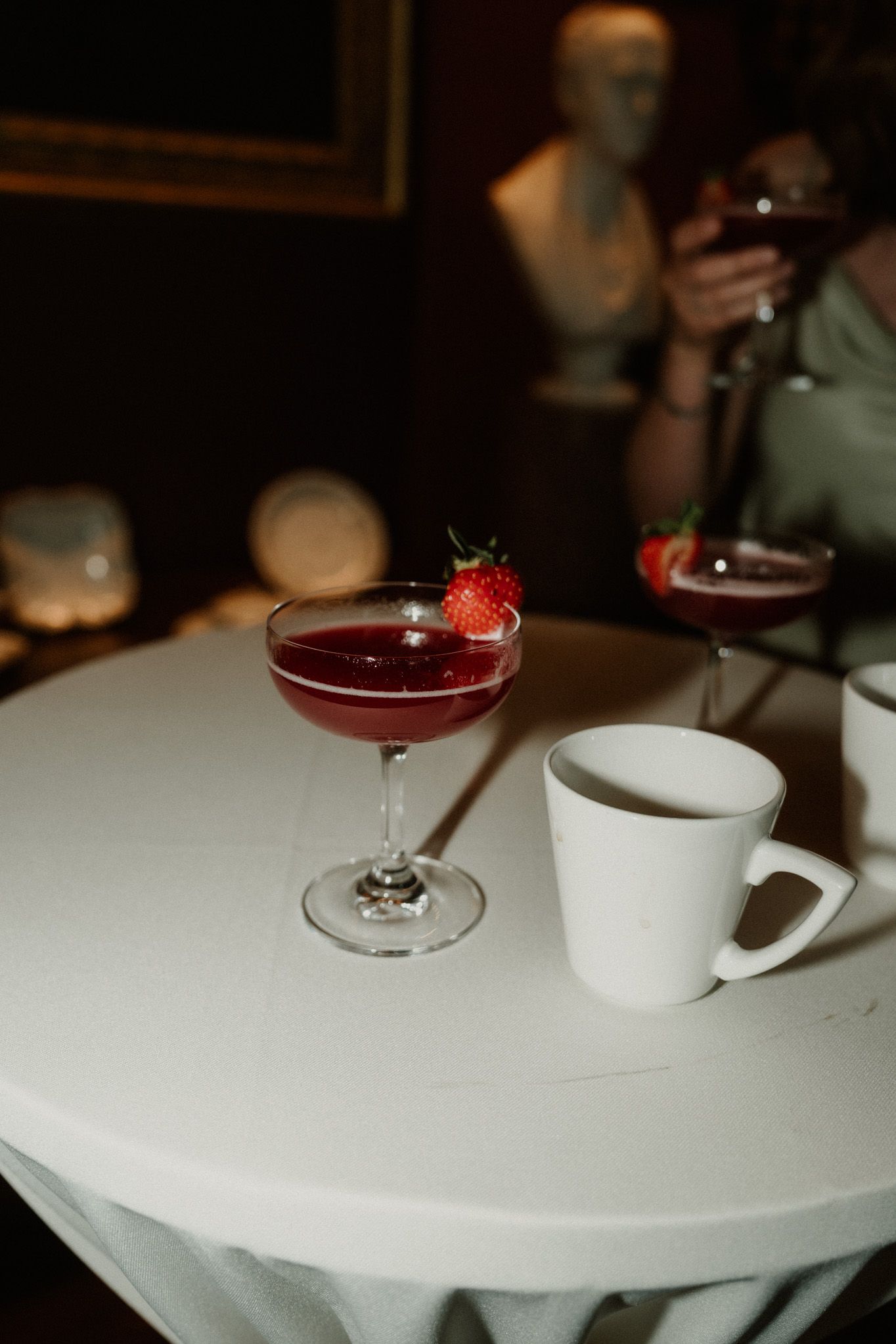 Strawberry-garnished red cocktail in a coupe glass beside a white mug on a round table, with a person holding a drink in the background.