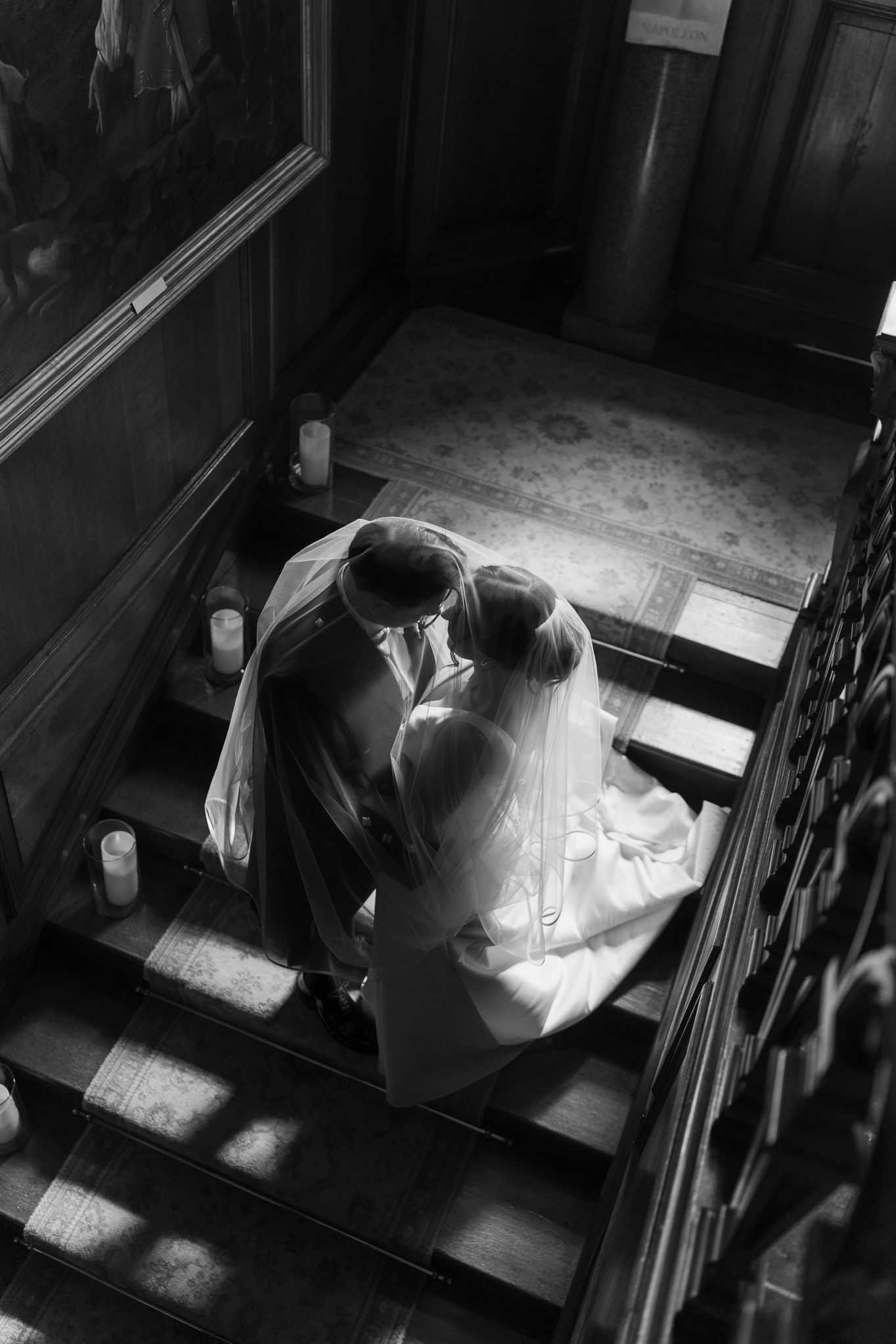 Black and white photo of a bride and groom embracing on a grand wooden staircase lined with candles in a historic venue.
