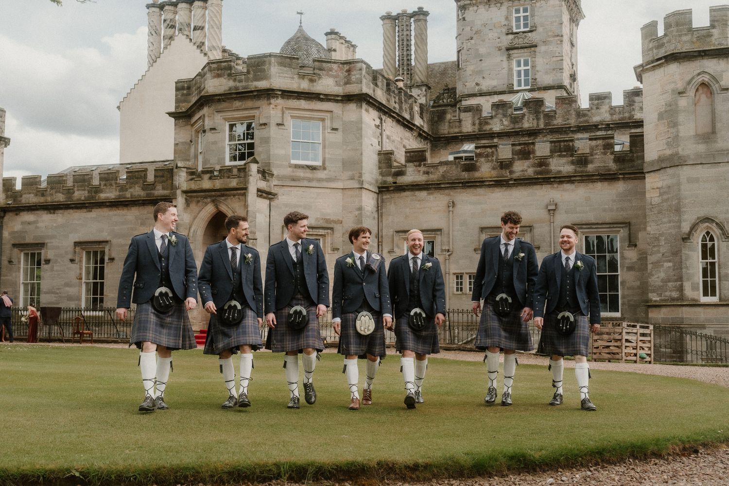 Seven groomsmen in kilts and dark jackets walk together outside a grand Scottish castle on a wedding day.