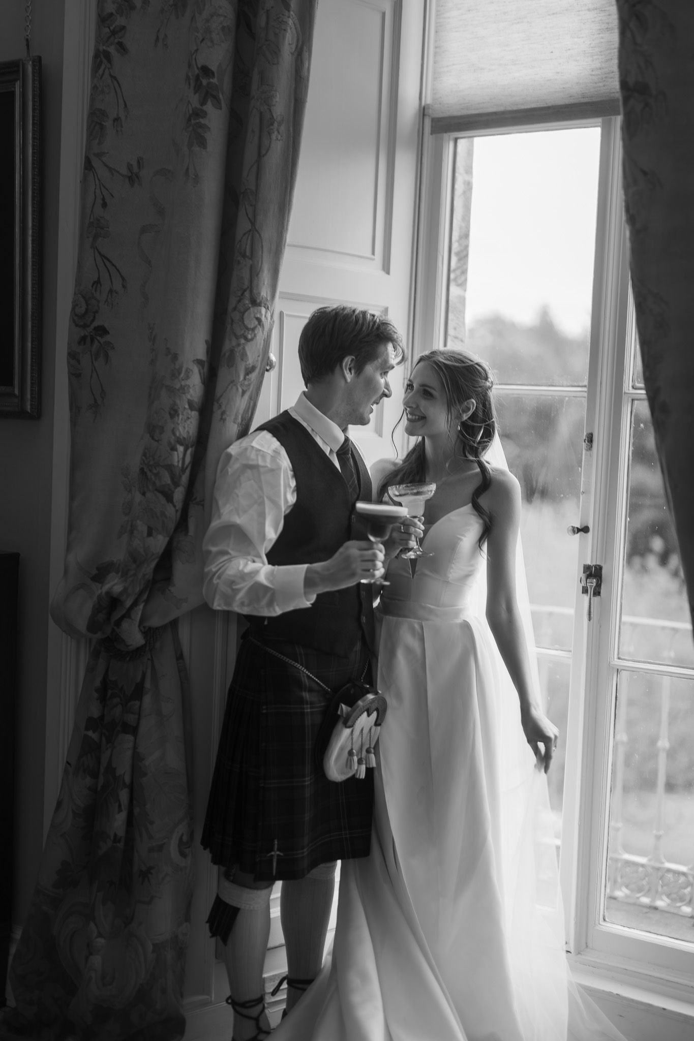 Black and white photo of a bride and groom in Scottish kilt sharing a romantic moment by an open window.