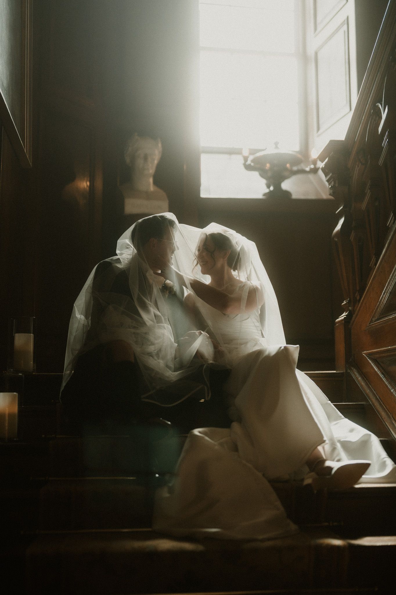Bride and groom share an intimate moment under a sheer veil on a wooden staircase, lit by soft window light beside a classical bust.