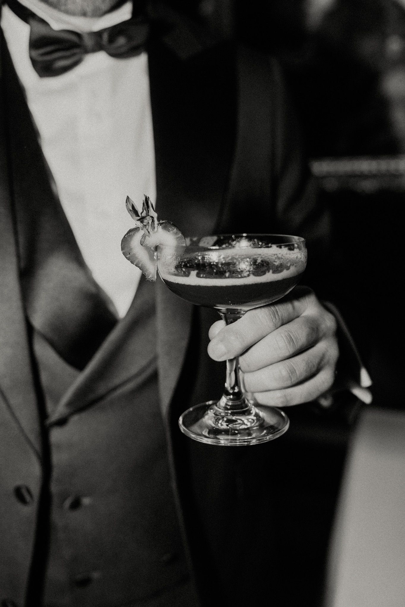 Black and white photo of a man in a tuxedo and bow tie holding a coupe cocktail garnished with a strawberry.