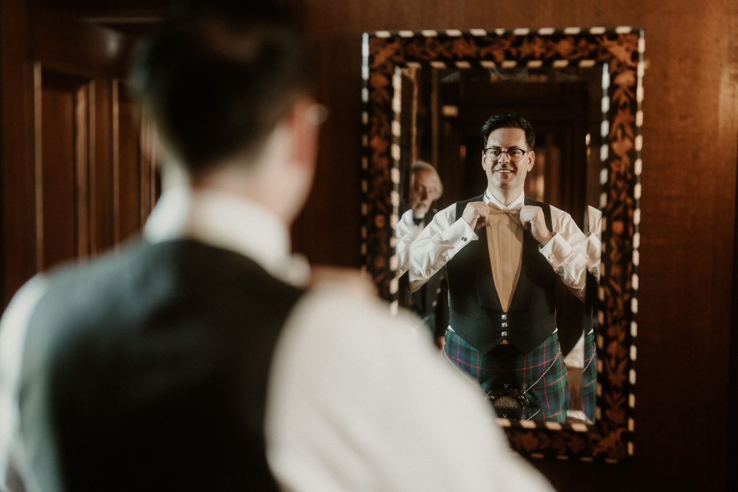 Groom in a vest and kilt adjusts his shirt cuffs while looking at his reflection in an ornate mirror in a warmly lit wood-paneled room.
