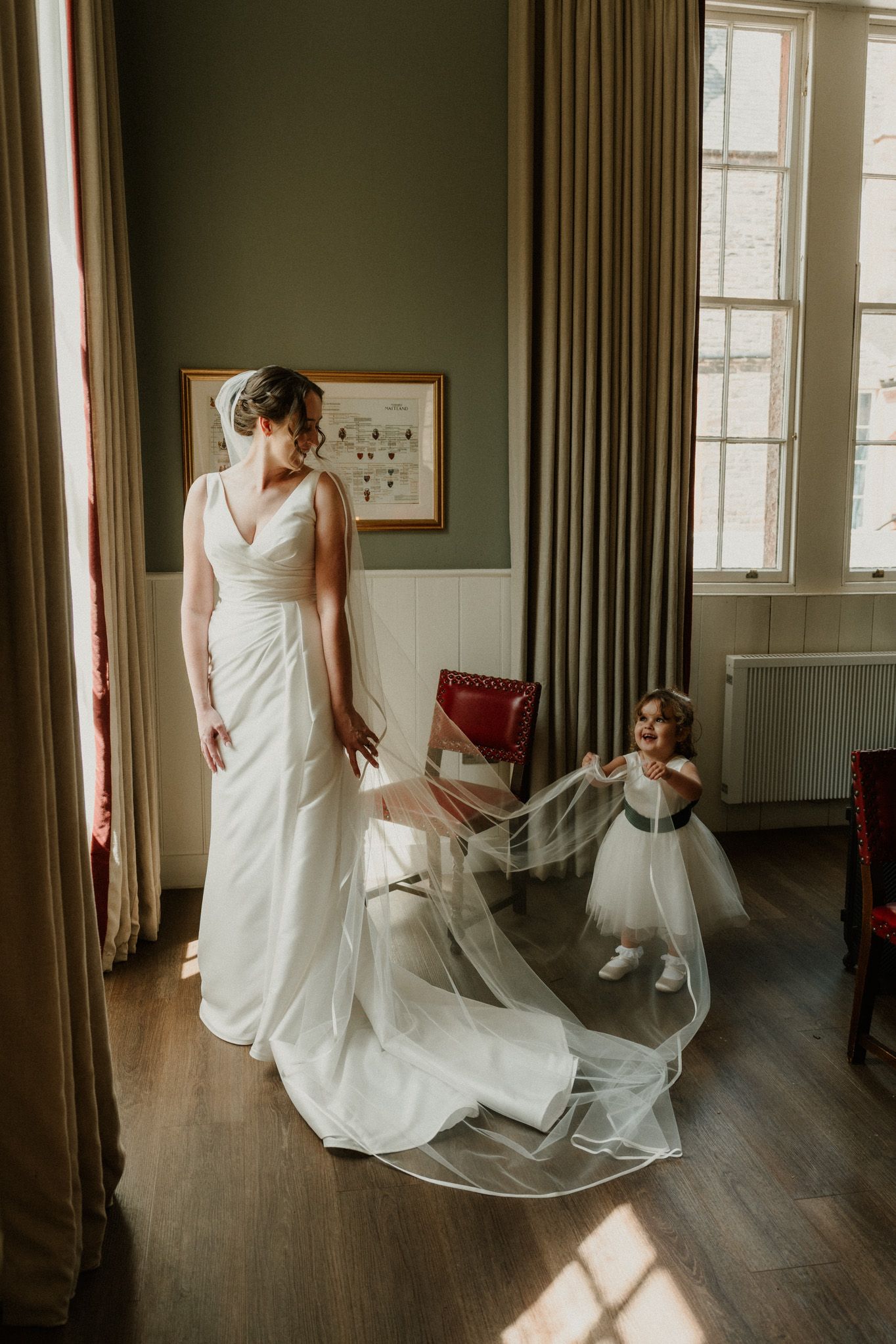 Bride in an elegant white gown with a long sheer veil stands by a window while a smiling flower girl in a tulle dress plays with the veil.