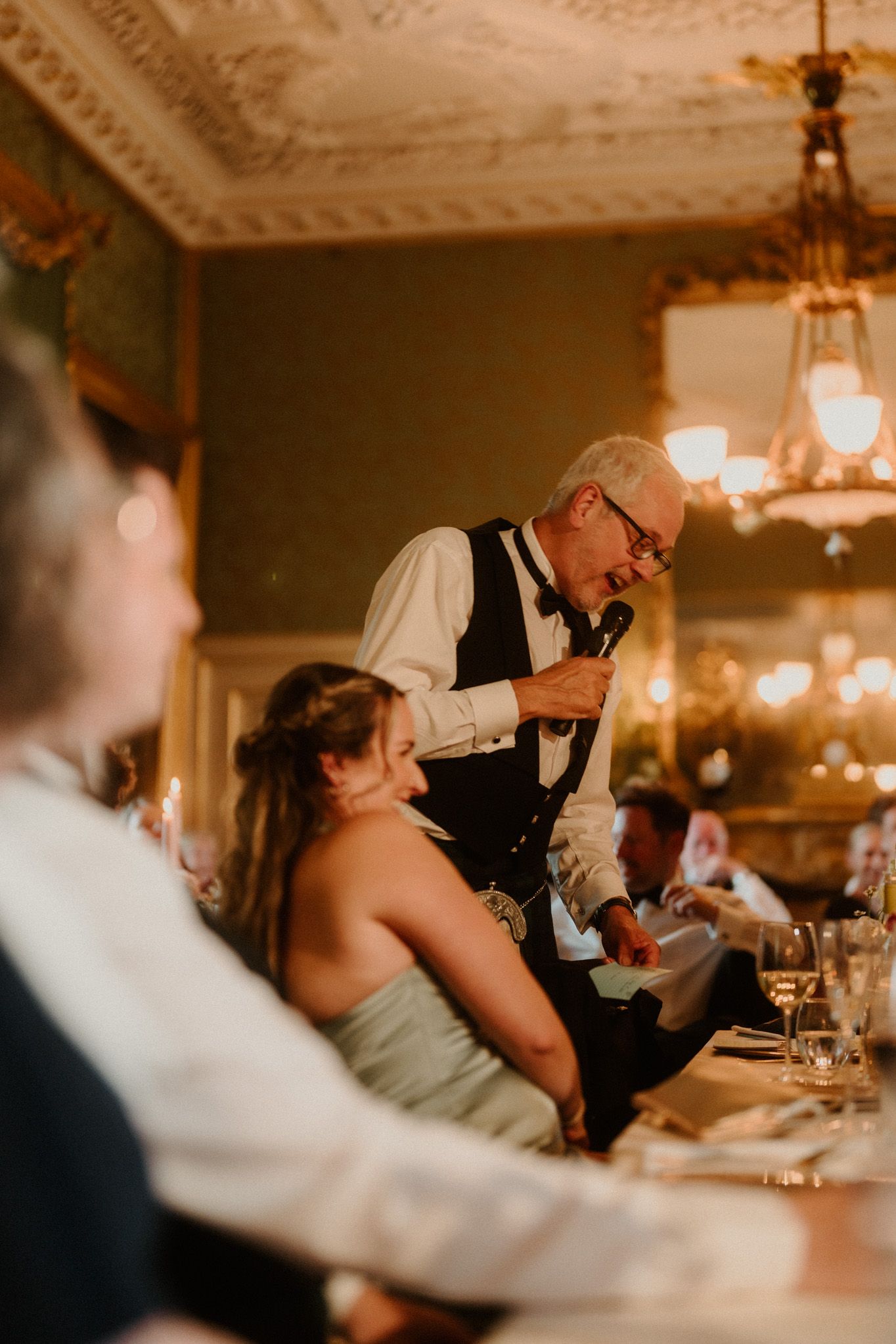Older man in a vest and bow tie gives a speech with a microphone at an elegant wedding reception while a seated woman listens.