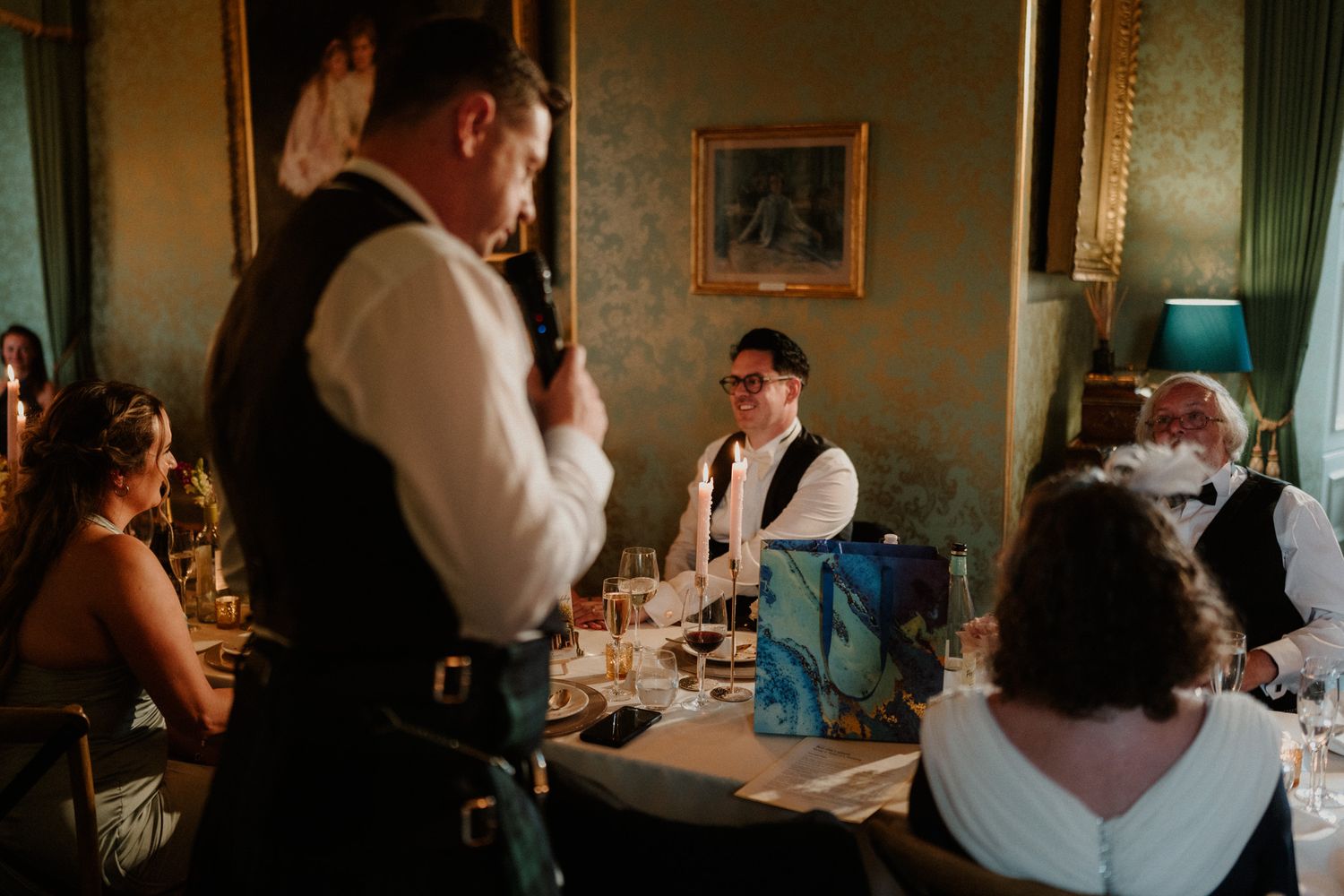 A man in a vest and kilt speaks into a microphone to seated wedding guests at an elegant candlelit dinner table in a formal room.