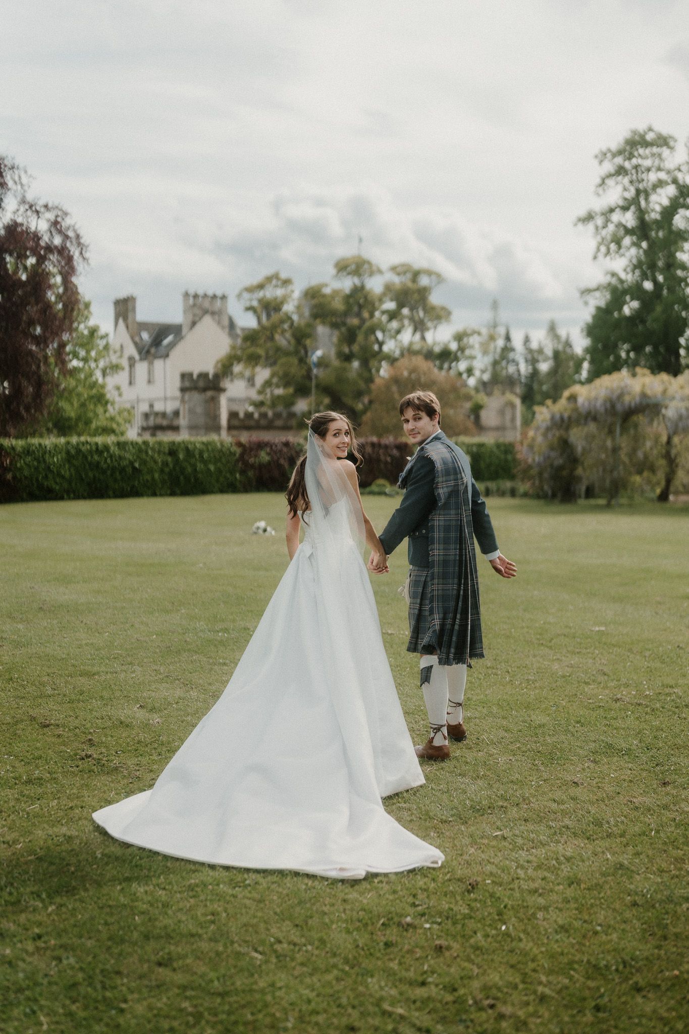 Bride in white gown and groom in Scottish tartan kilt hold hands on lush green lawn with historic manor backdrop.