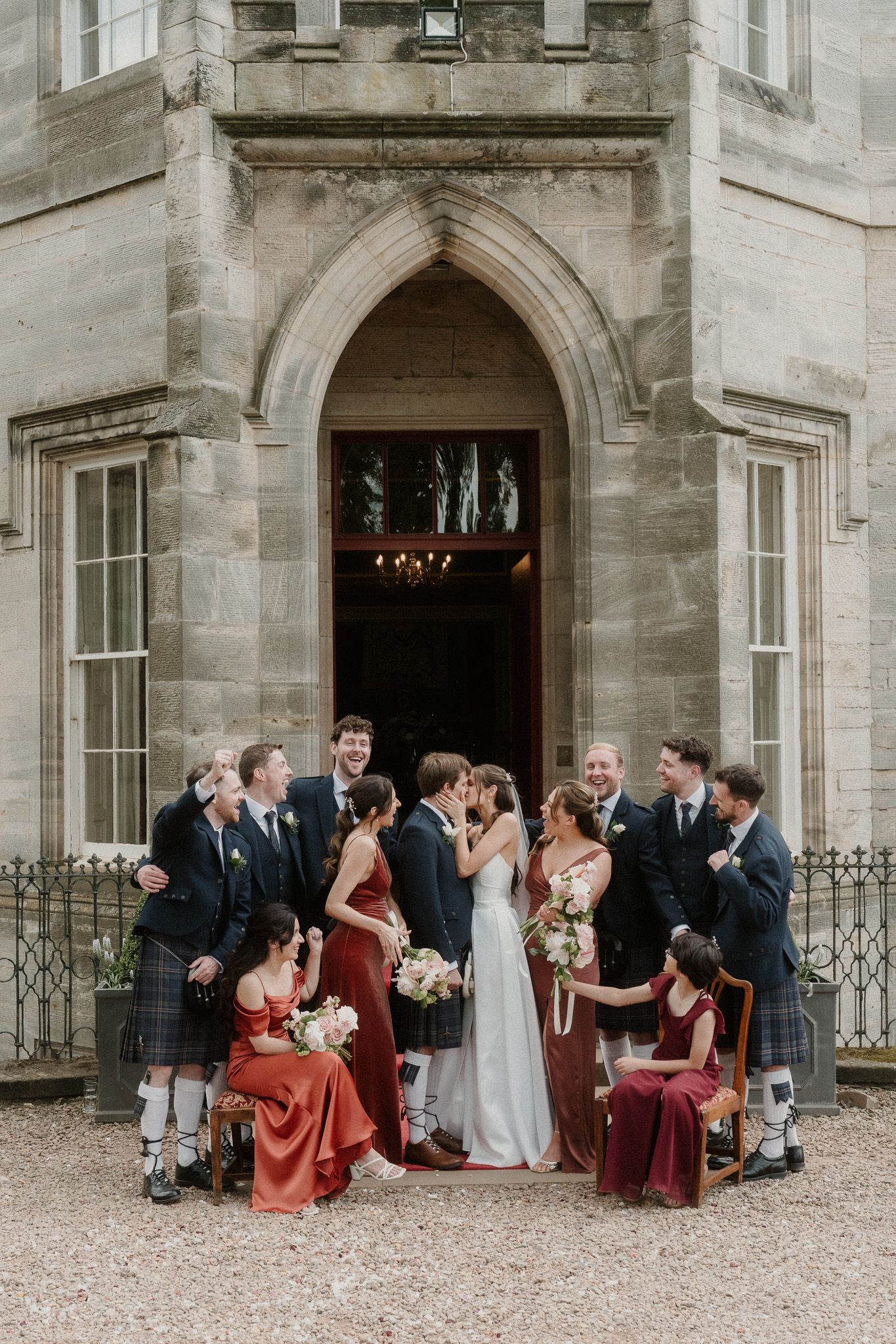 Wedding party kisses outside a Scottish castle, bridesmaids in red dresses and groomsmen in kilts surrounding the couple.
