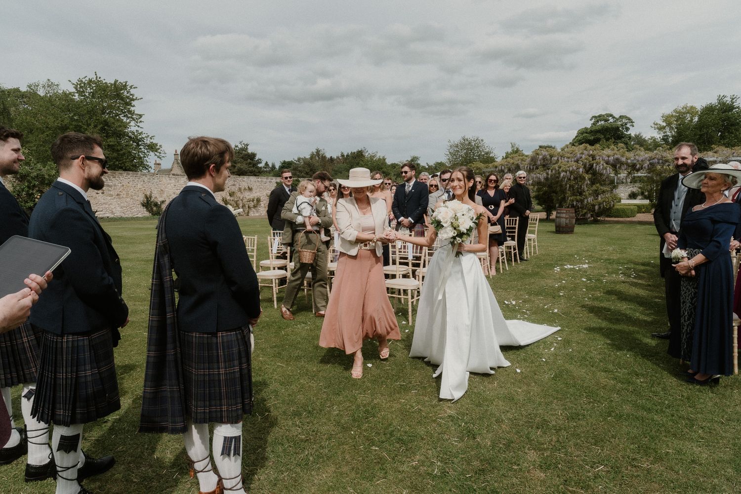 Bride walking down outdoor aisle with escort at elegant garden wedding ceremony, guests watching in Scottish attire.