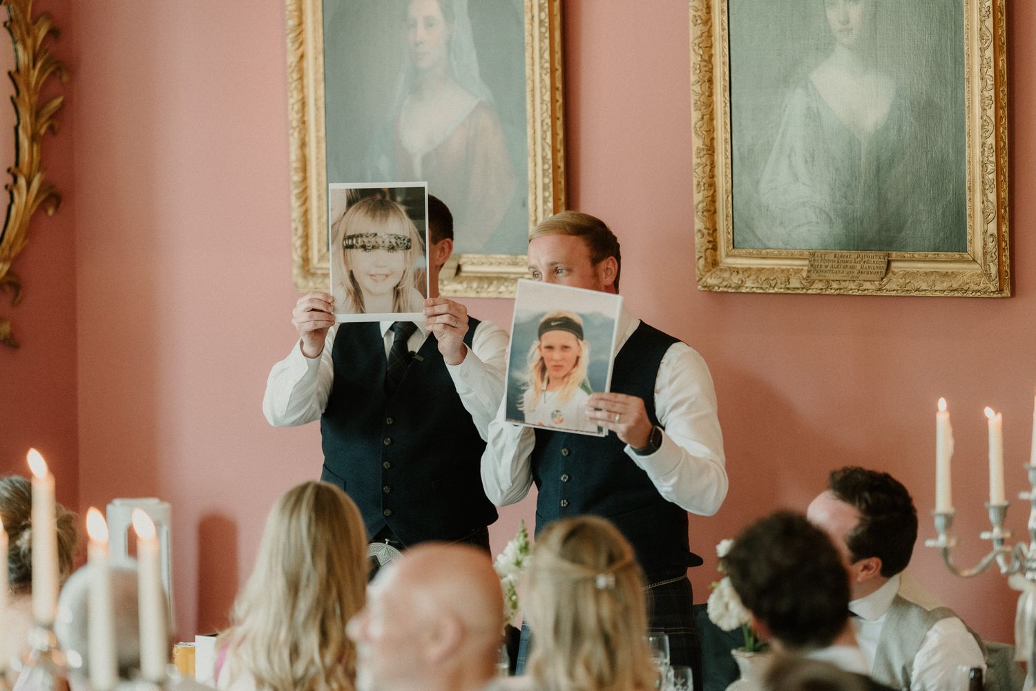 Two groomsmen holding up childhood photos of the bride and groom during a wedding speech in an elegant room.