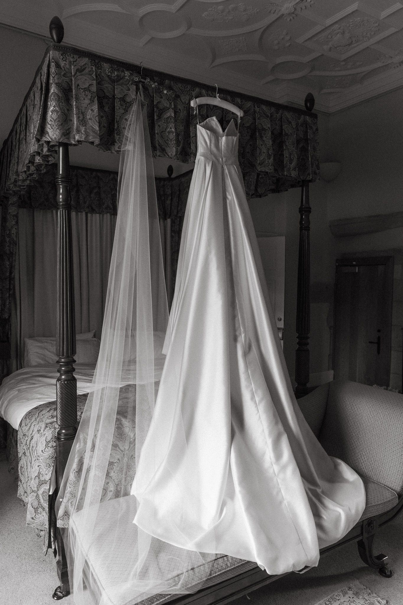 Elegant wedding dress with veil hanging on a four-poster bed in a grand bedroom, black and white photography.