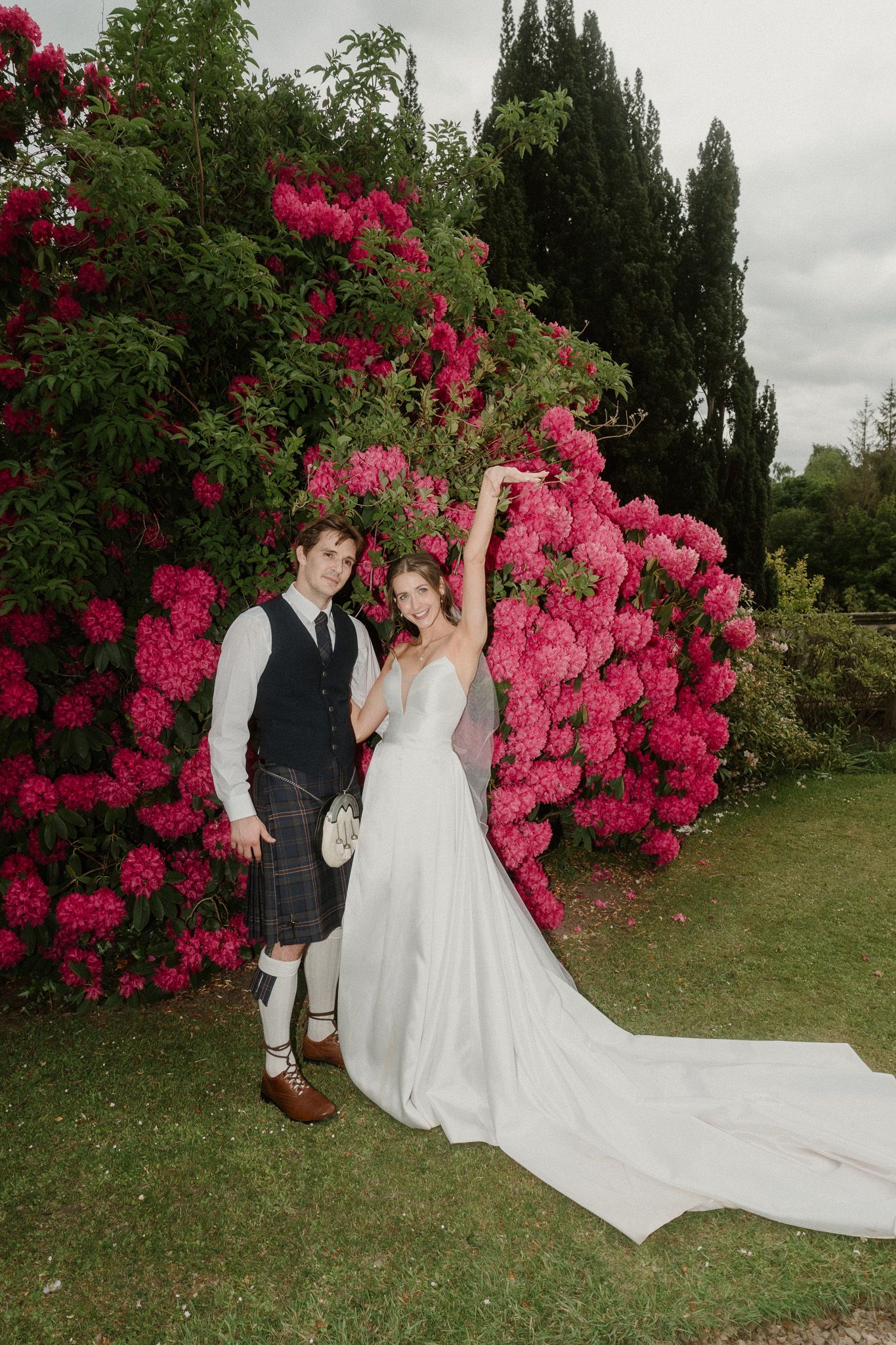 Bride in white gown and groom in Scottish kilt pose joyfully before vibrant pink rhododendron bushes in lush garden.