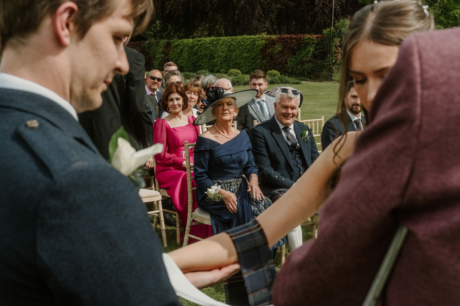 Outdoor wedding ceremony with guests smiling as couple exchanges vows, woman in blue dress stands prominently among attendees.