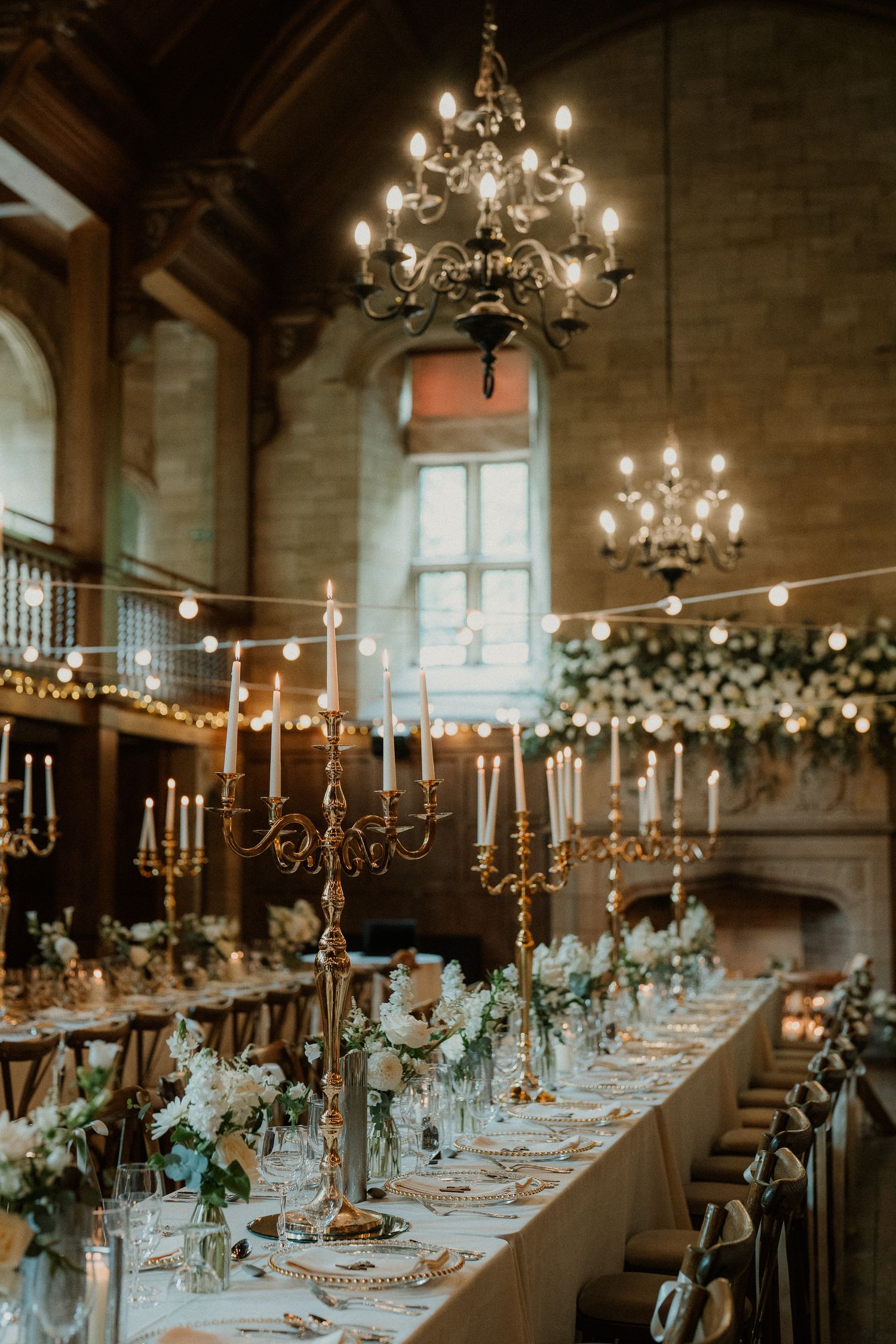 Elegant wedding reception in a grand hall with candelabras, chandeliers, floral arrangements, and a long dining table set for guests.